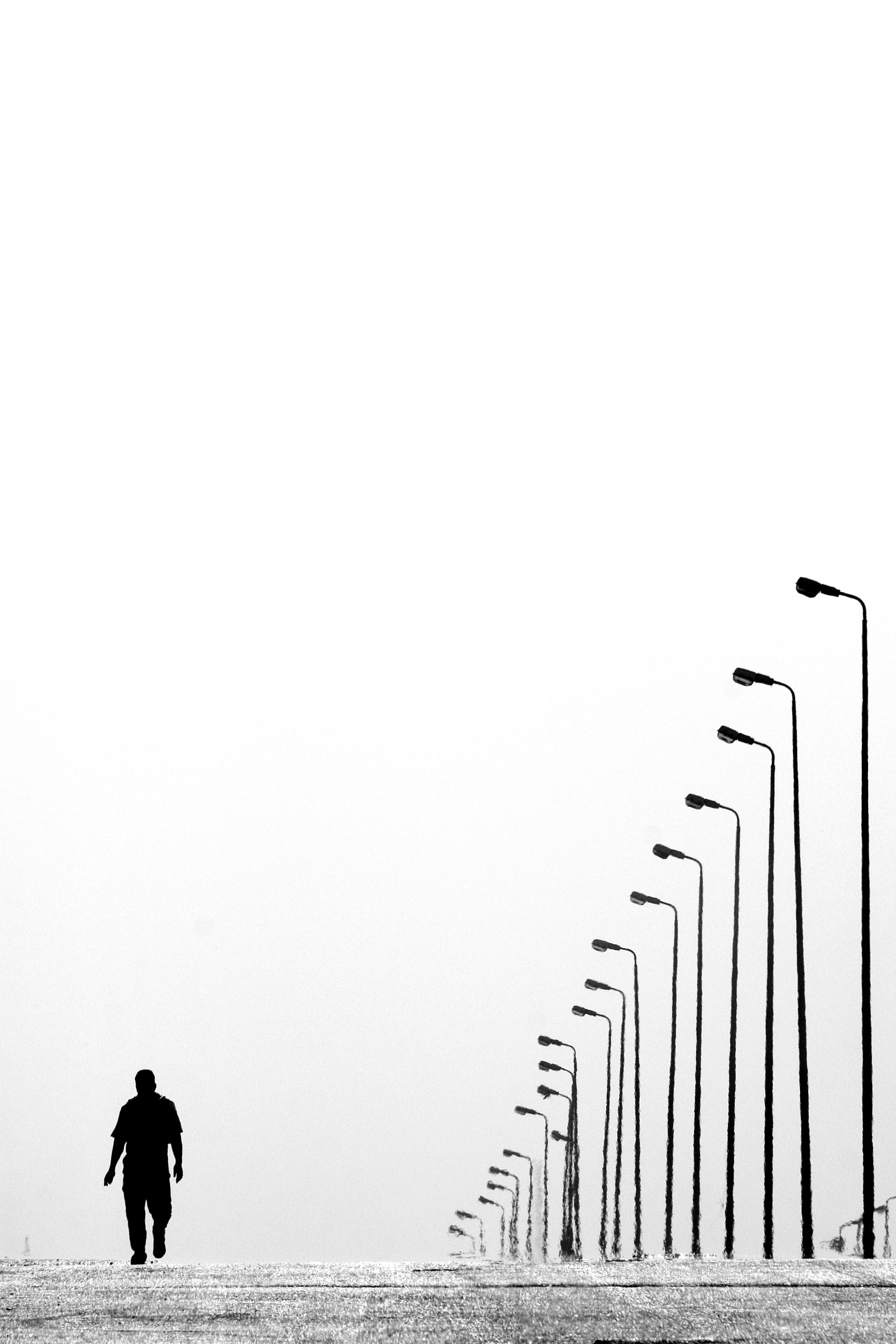 Black and White photo of man walking on empty road alongside lights