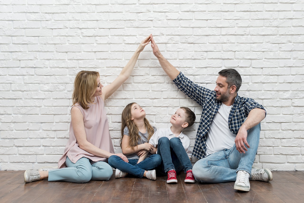 Two children sitting below and looking up at Mom and Dads hands touching forming a protective arch as they all sit on the ground against a white brick wall.