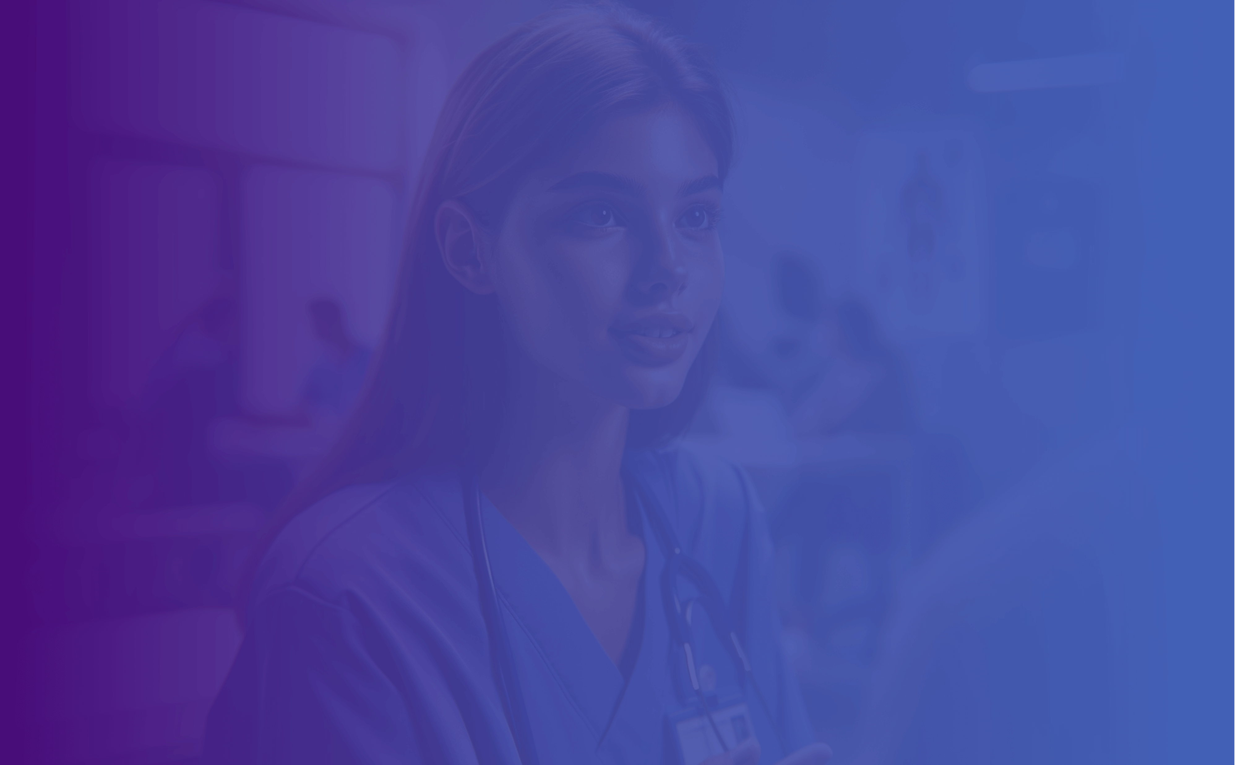 A young female medical professional in a blue scrubs uniform is attentively listening to someone off-camera, with a hospital setting and other medical staff in the blurred background.