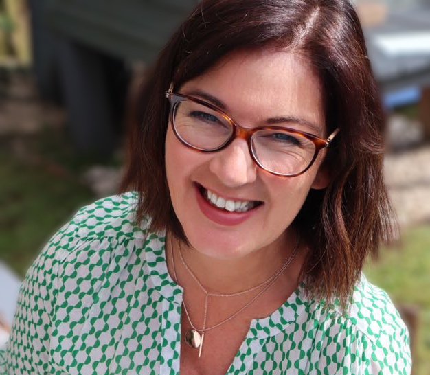 A photo of Tara Killen, a smiling white woman with brown hair wearing glasses outdoors in the sunshine.