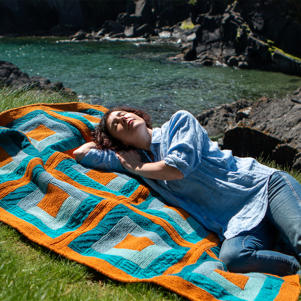 Wom,an lying down on a larage colourful log cabin style blanket with her eyes closed.