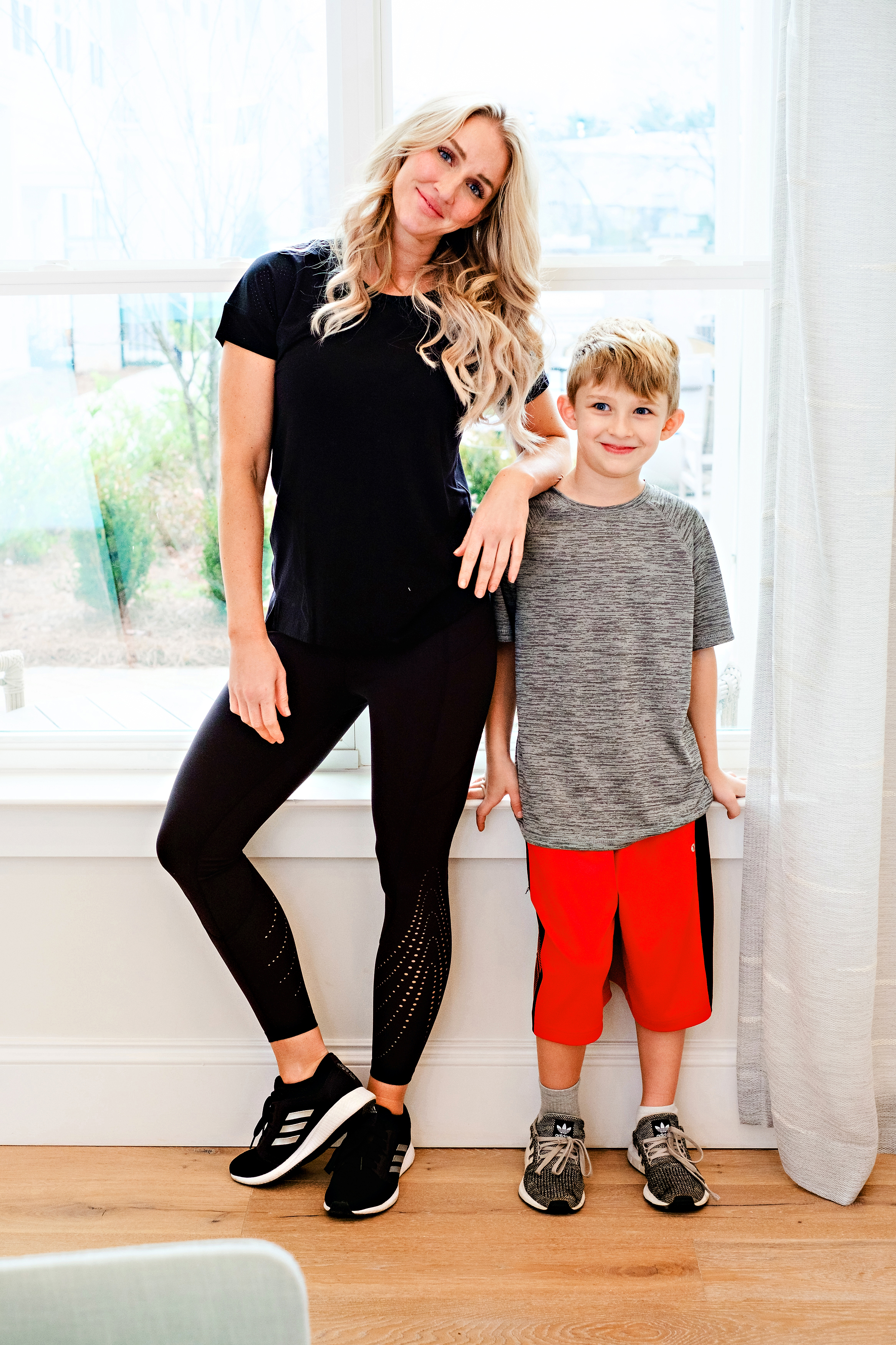 Blonde woman named Jessica from Happily Hughes in black leggings with a black t-shirt leaning her arm against her son who is in a grey t-shirt and red shorts smiling for the camera!