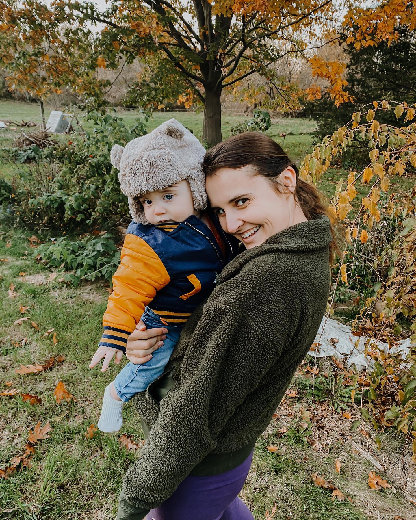Jennifer in a green jacket and purple leggings with her 1yr. old son Malachi, standing outside in the garden