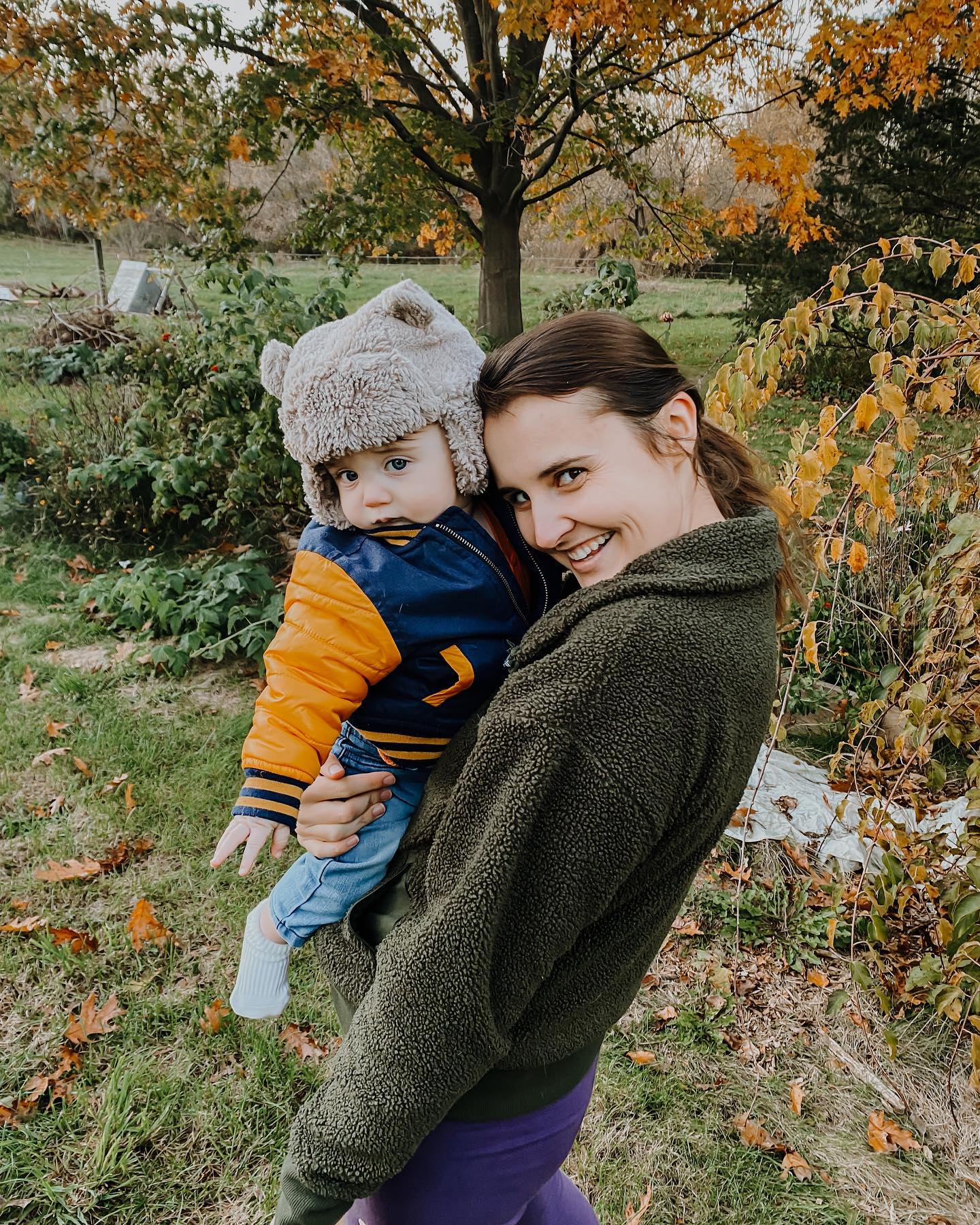 Jennifer in a green jacket and purple leggings with her 1yr. old son Malachi, standing outside in the garden