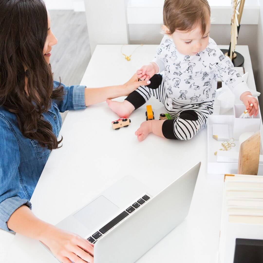 A woman working at a computer with her baby on the desk