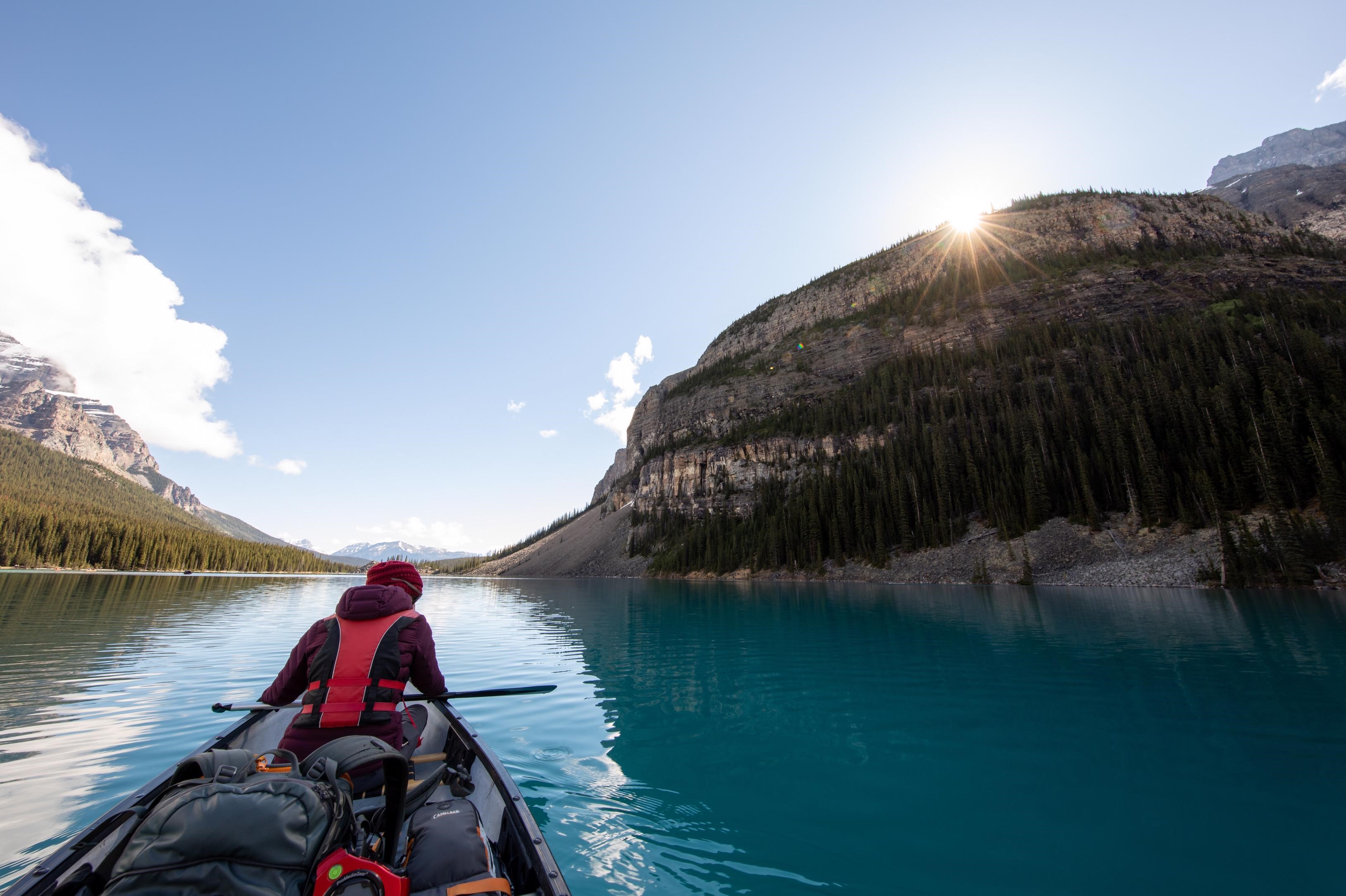 A person sits in a canoe on a bright blue lake next to a large mountain, the sun shining in the background.