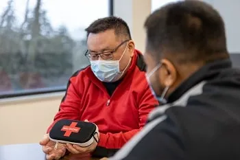 A person is holding and showing a Naloxone kit to another person, both wearing masks.