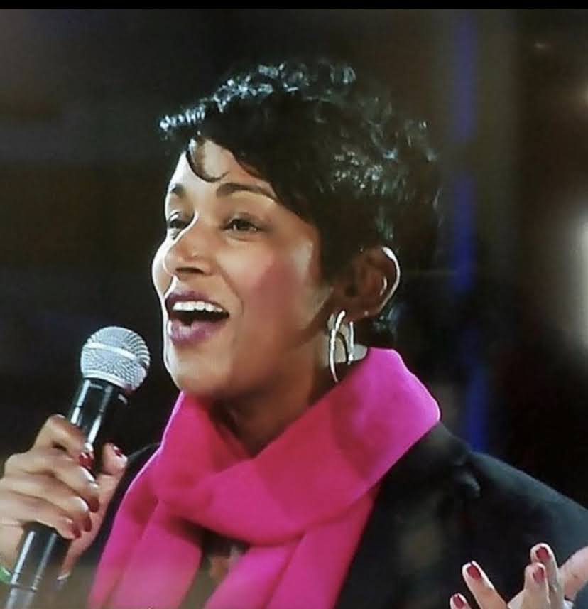 image of Leatha, speaking into a microphone, wearing a pink scarf