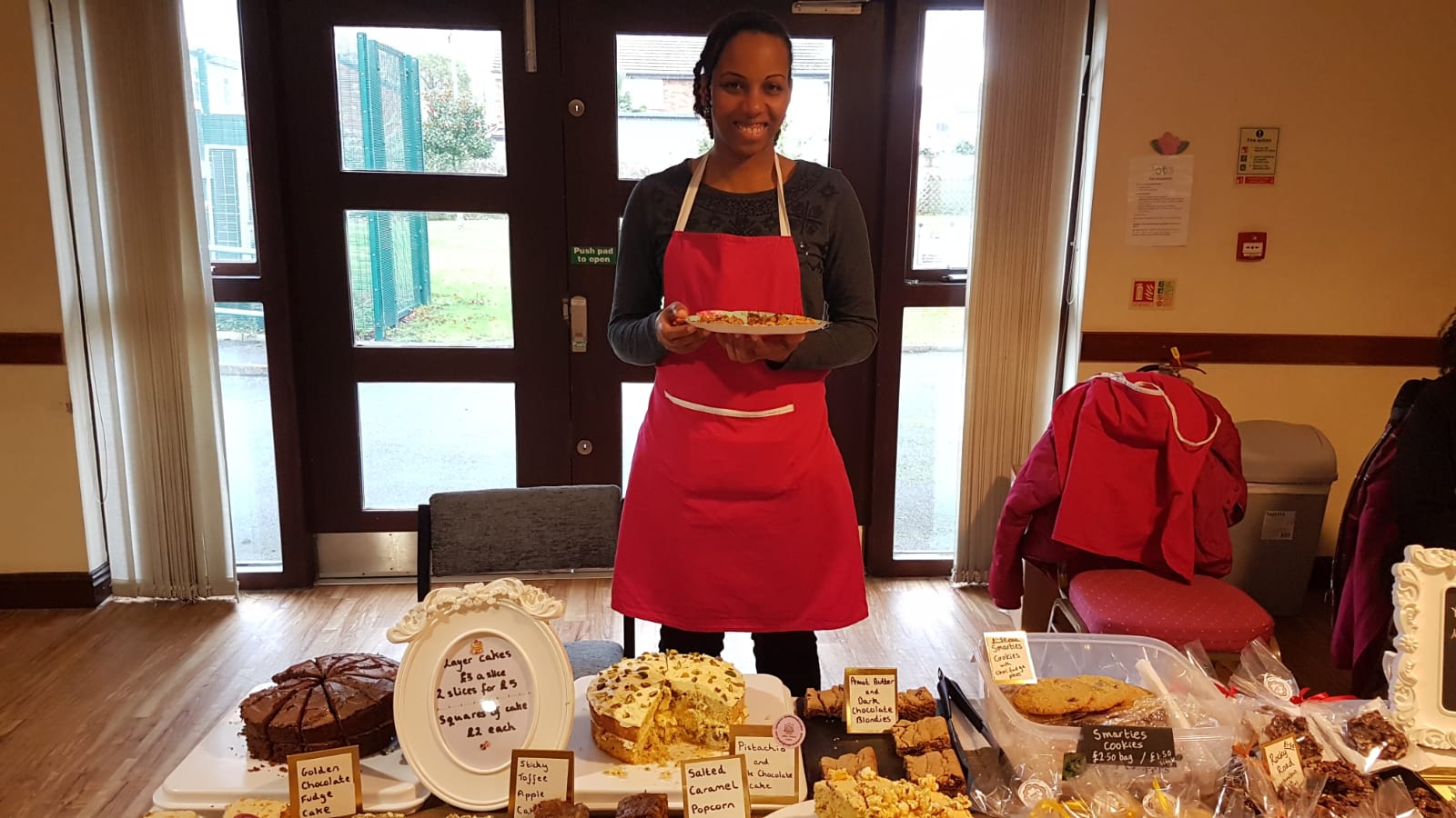 A female wearing an apron is standing behind a table of cakes and cookies. She is holding a plate of cakes.