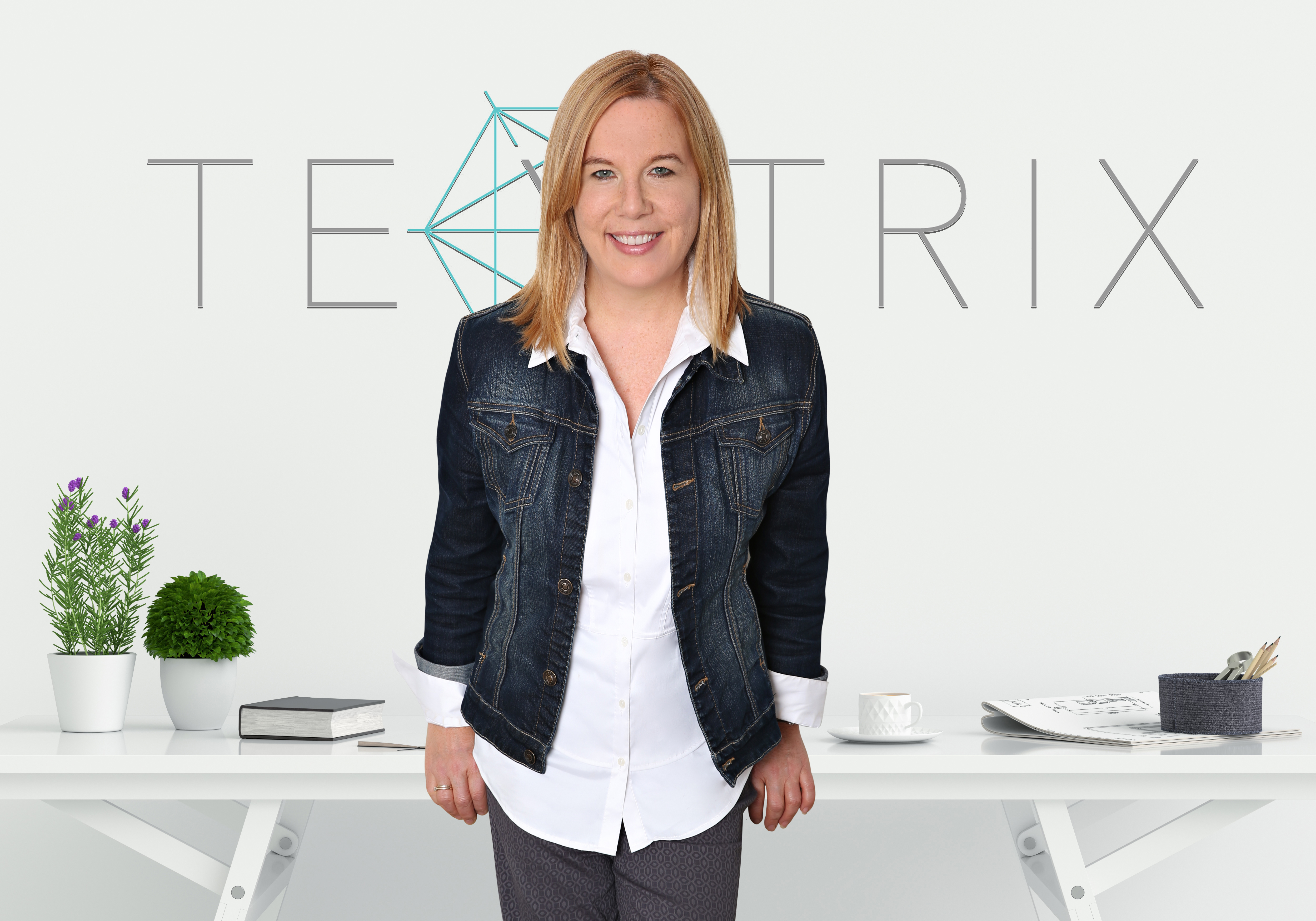 A woman with shoulder length red/blond hair stands in front of a table wearing a white shirt and denim jacket. She is smiling