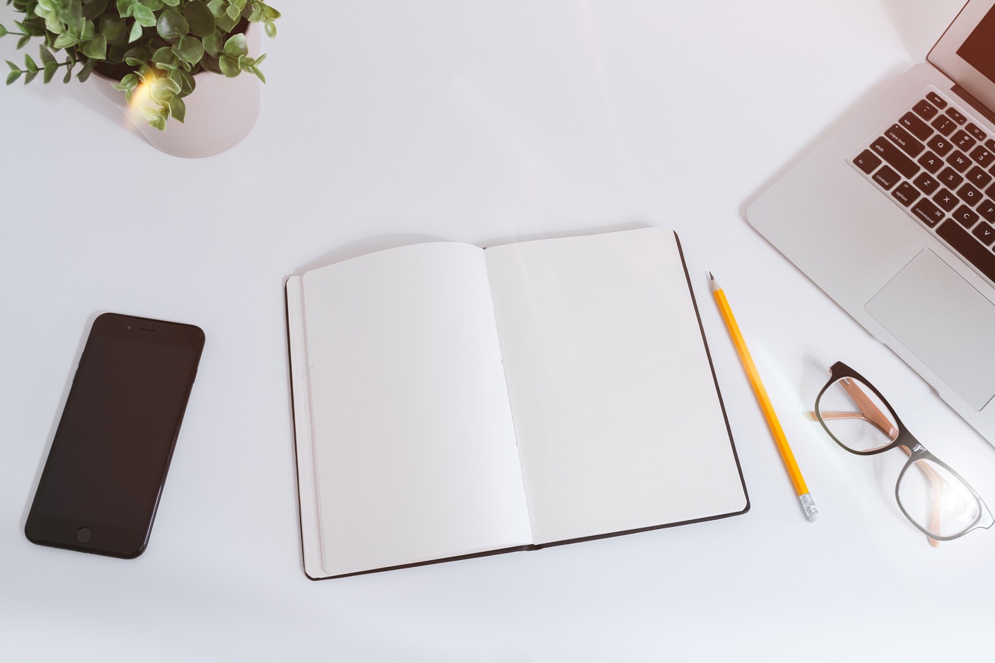 Photo. Open notebook lying on desk. Pencil, eyeglasses, and part of open laptop at right. Cell phone at left. Part of small plant upper left corner.