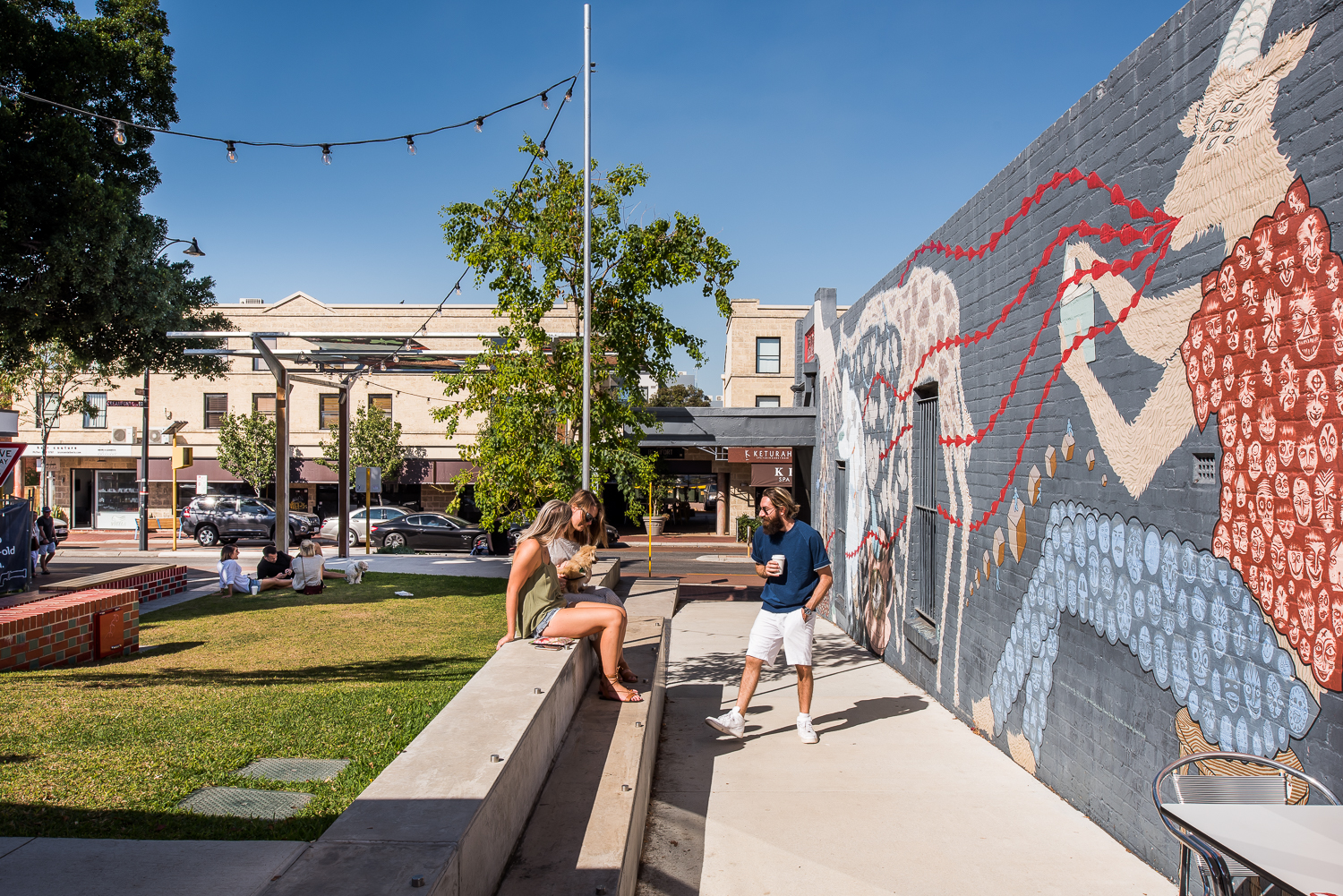 Picture shpwing Mary Street piazza on Beaufort Street, Highgate, Western Australia. One man is drinking a coffee and two women are talking. A large mural by Rob Jenkins is shown on a wall to the right whilst a grassed area is to the left with Beaufort Street in the background