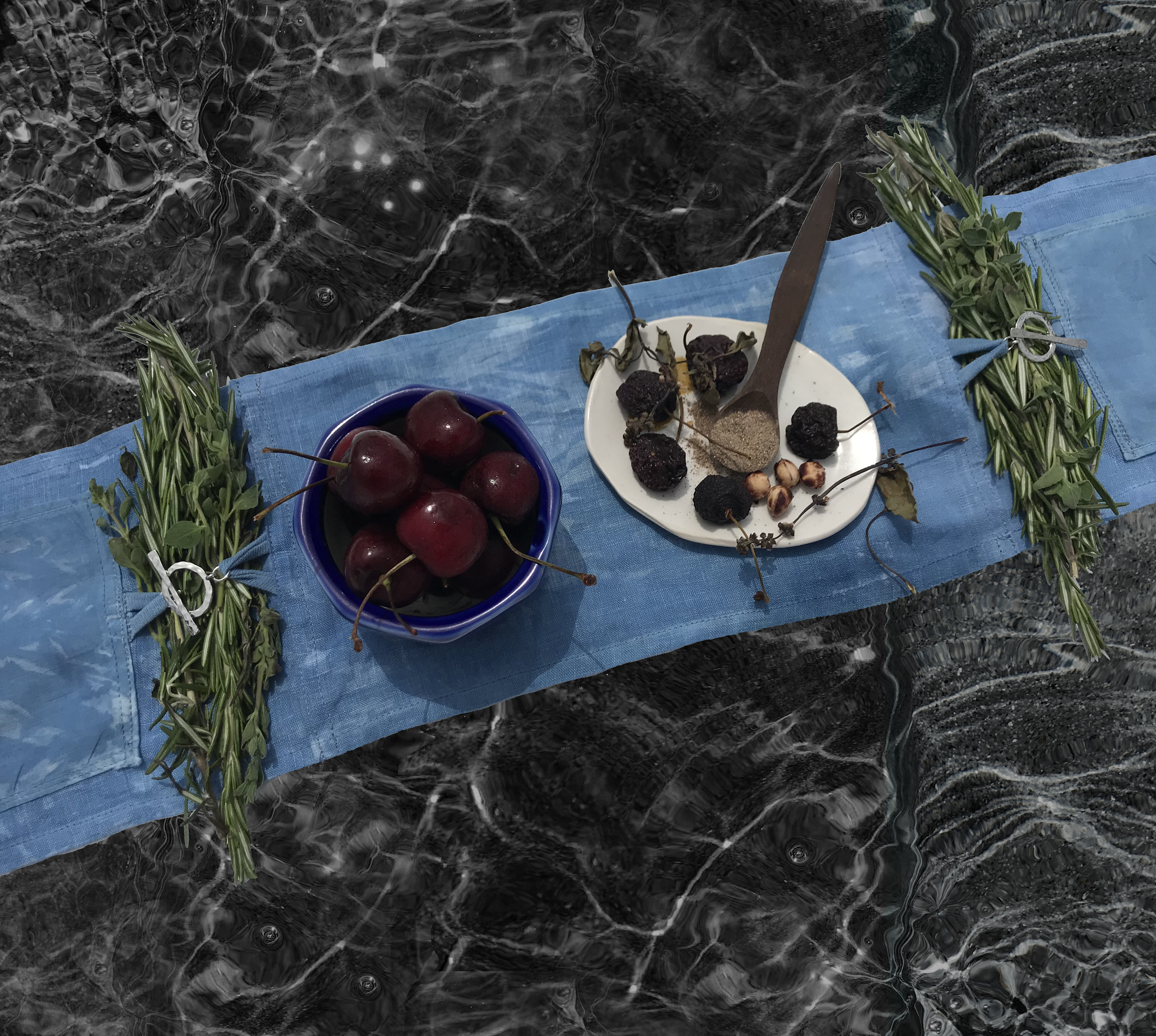 an indigo dipped tapestry hovers over a watery black background. on the tapestry are two clusters of rosemary, a bowl of fresh cherries and a dish of dried cherries with a spoon full of cardamom. 