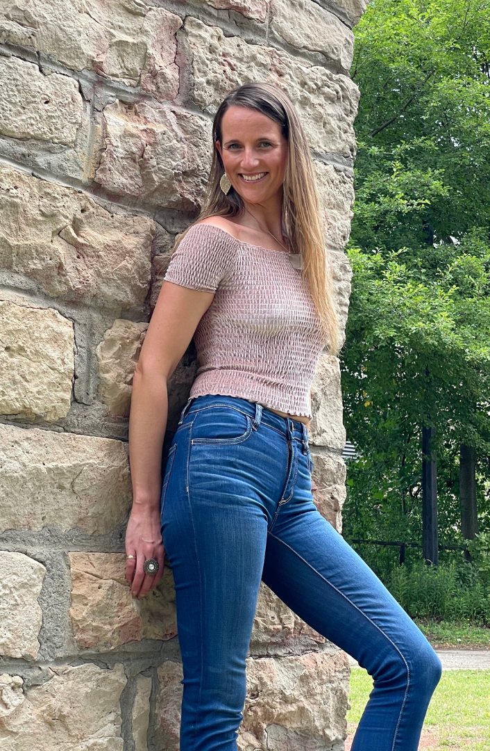 Woman smiling with her back leaning against a stone wall with green trees in the background
