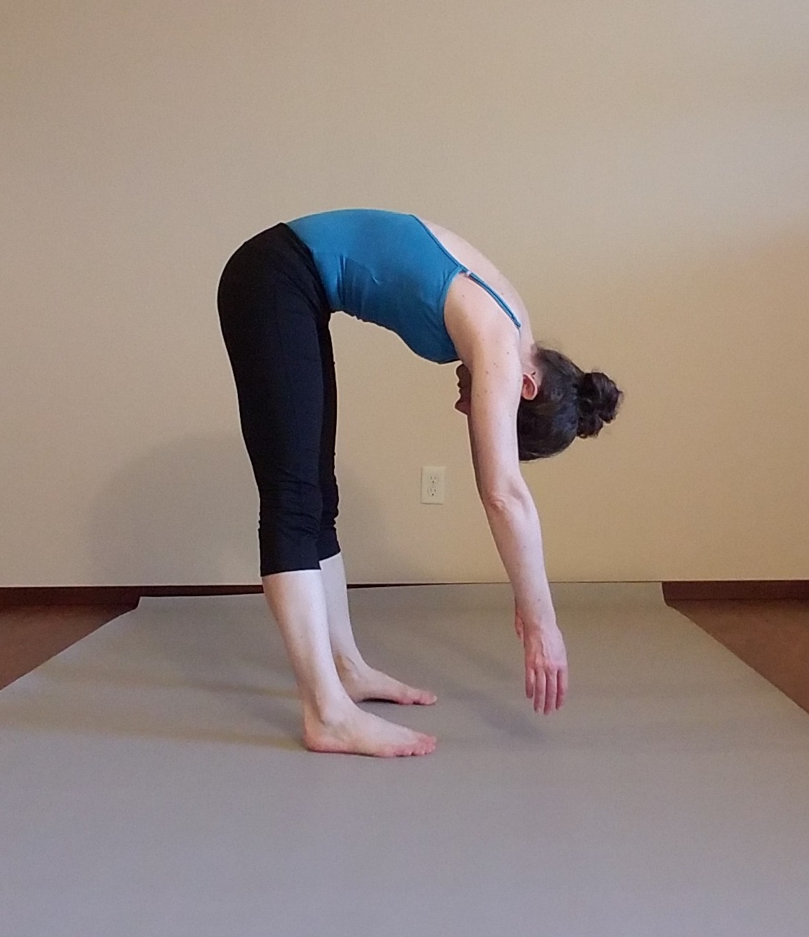 Instructor Kate Feinberg Robins demonstrates a Pilates Roll-Down standing on bare feet on a light gray floor at home with arms and head hanging down, back rounded and stomach scooped