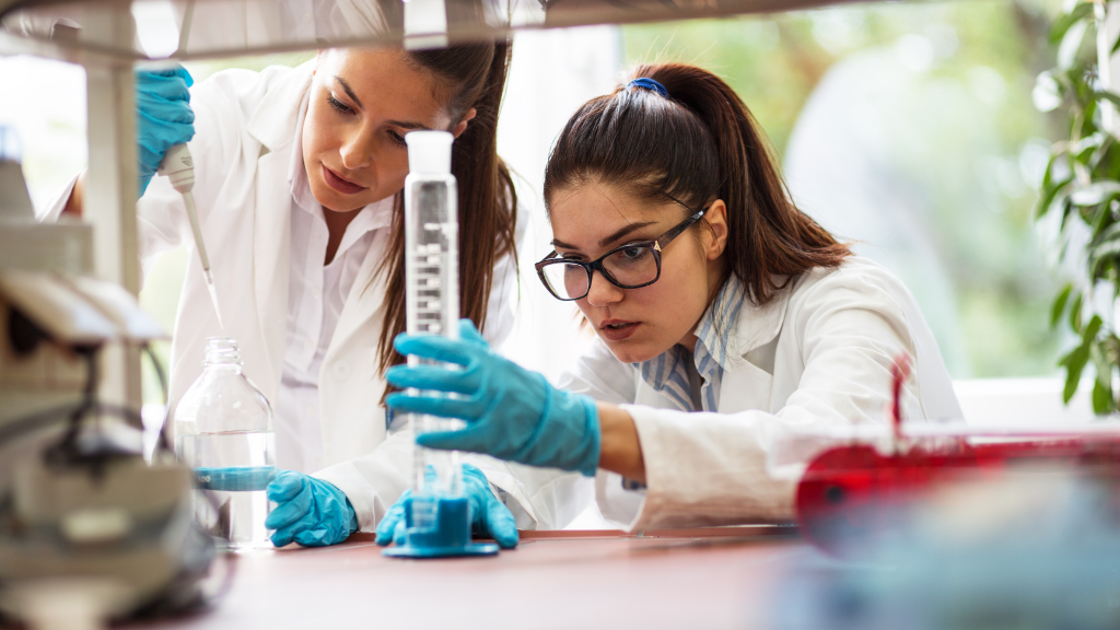Two young women in lab coats working together in a biology lab, using a pipette and graduated cylinder with scientific focus and collaboration.