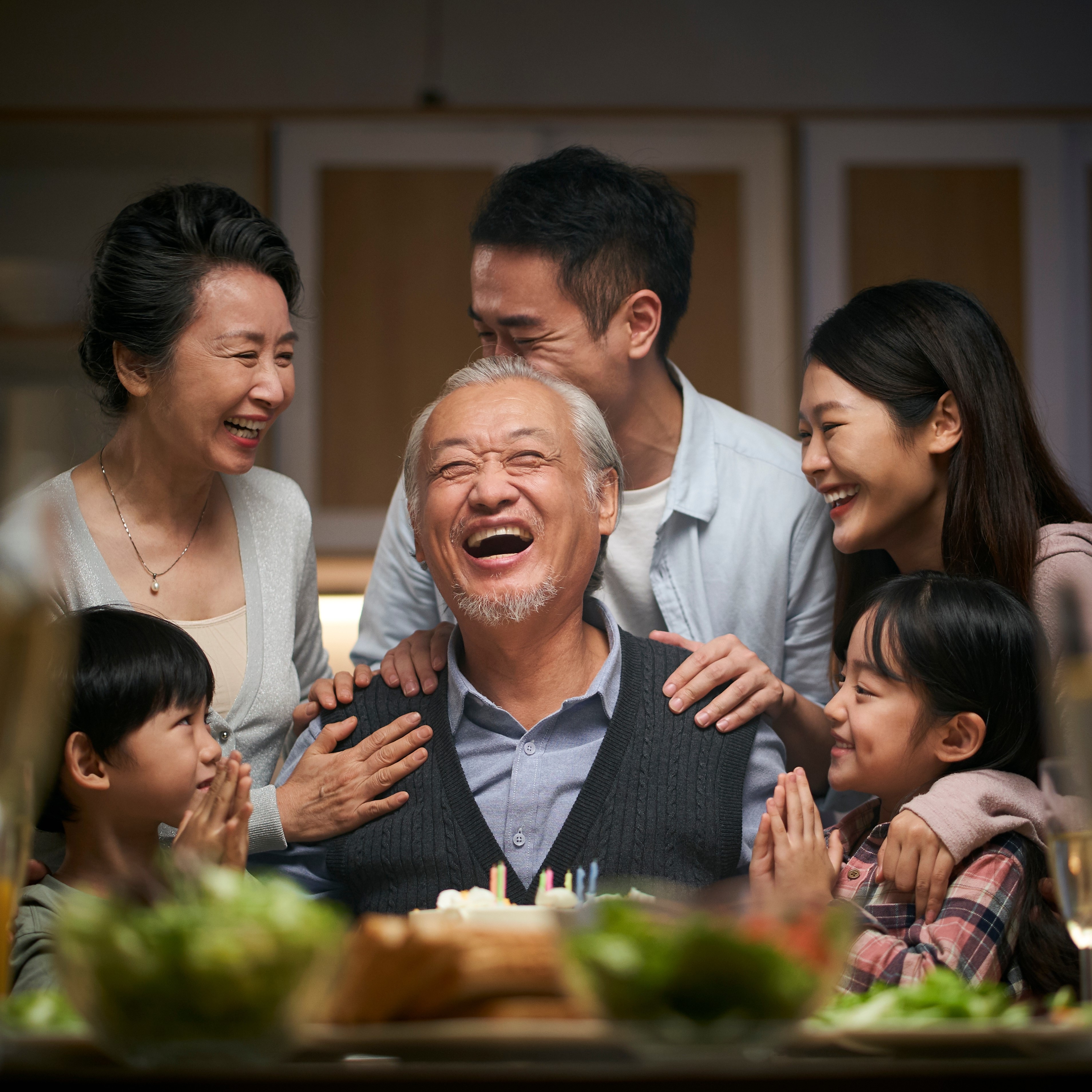 Retired man sitting at a table with a birthday cake in front of him, surrounded by his family, laughing