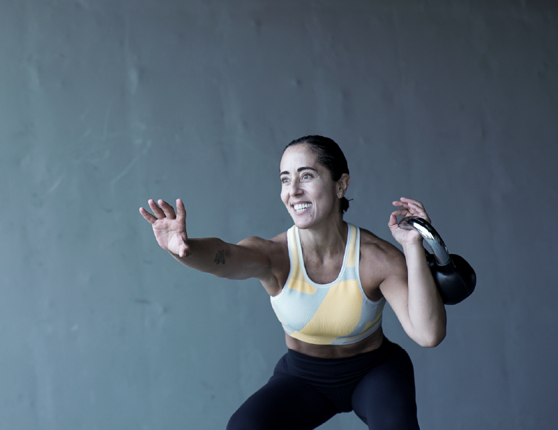 Mujer en ropa deportiva realiza ejercicios funcionales y de tonificación con un kettlebell en la mano izquierda.