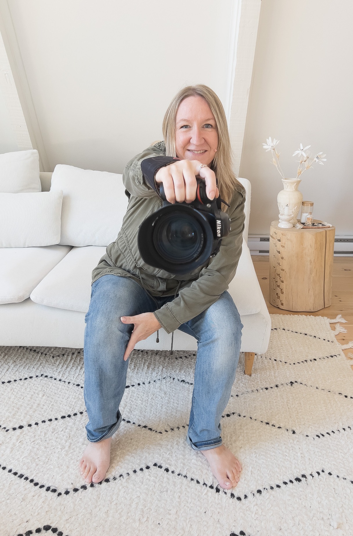 female photographer with blond hair, wearing olive green jacket and blue jeans, sitting on white sofa, holding out camera with large lens