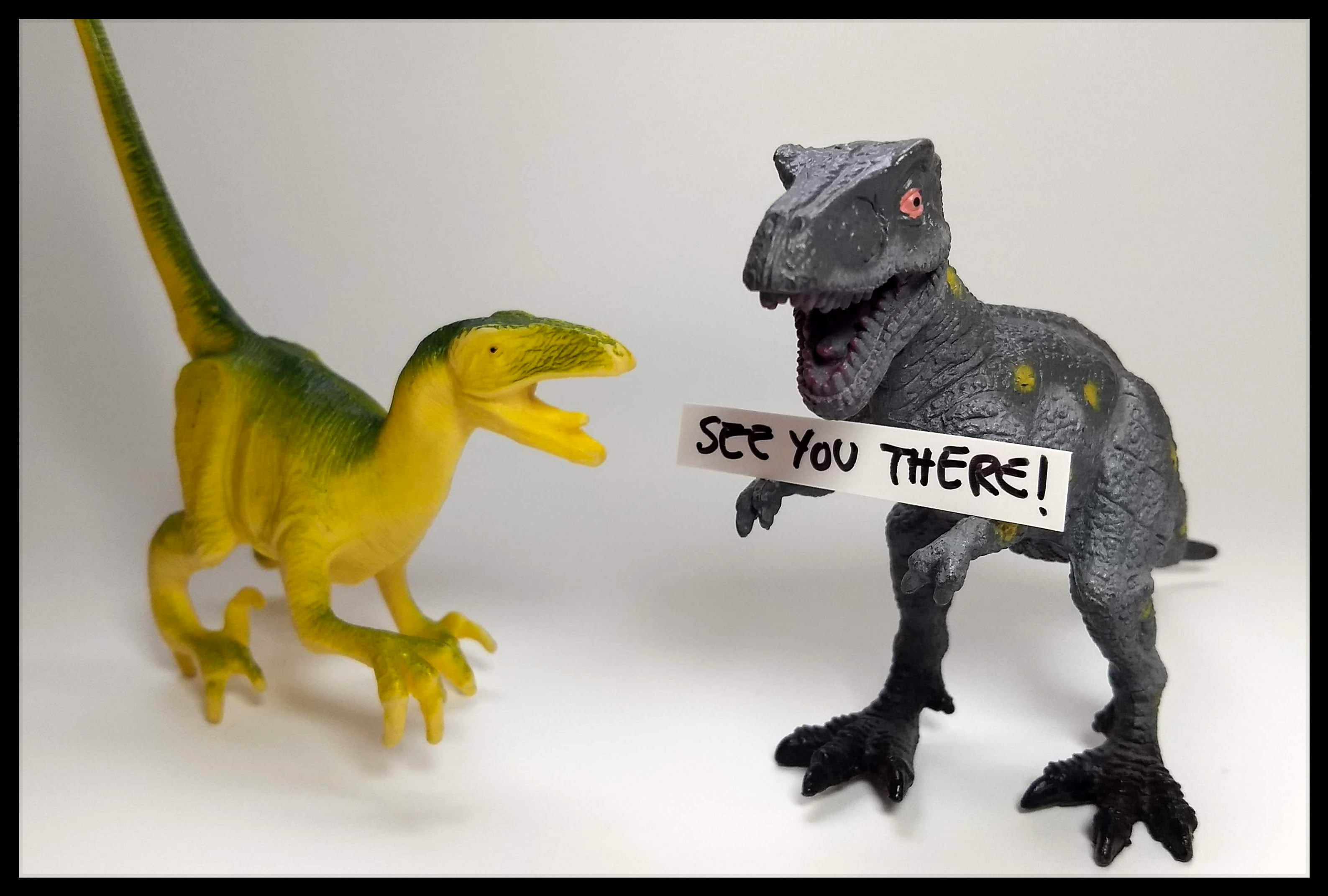Two toy dinosaurs. One is holding a sign that says SEE YOU THERE!