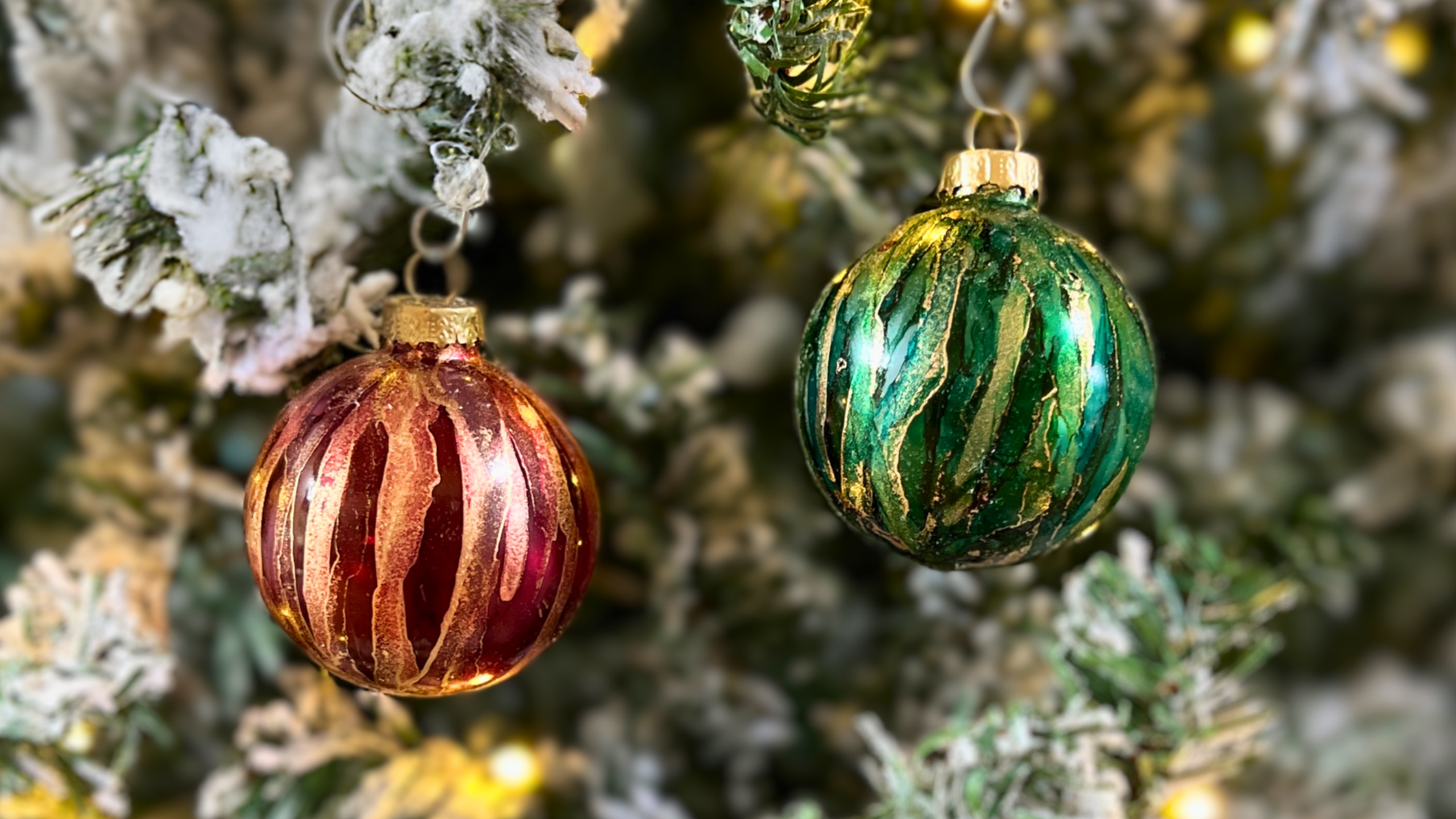 Handcrafted alcohol ink Christmas ornaments in red and green hanging on a snow-frosted tree, showcasing the artistry taught in our online course.