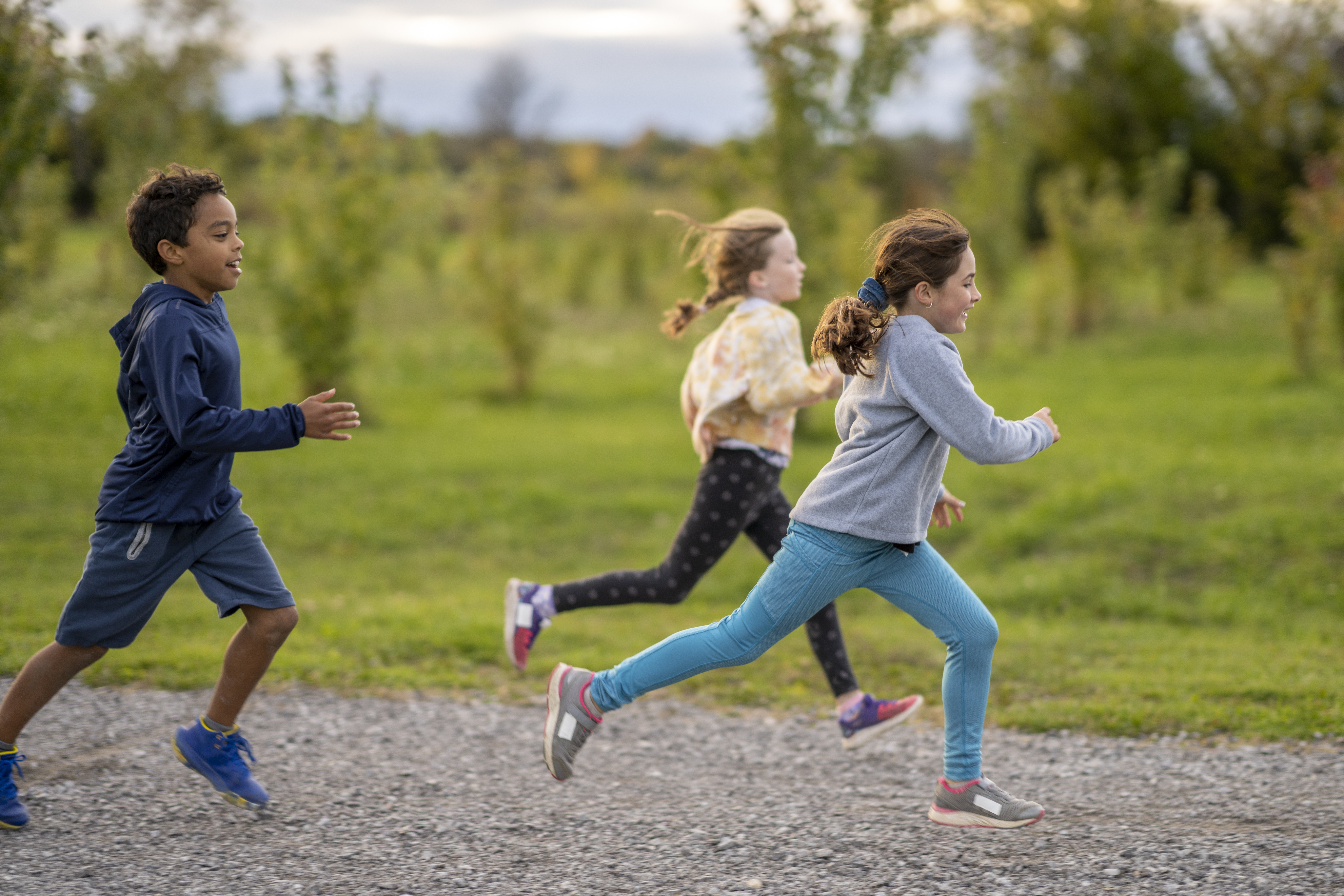 Photo of three elementary-school students running on a gravel track from right to left. In the background are grass and small trees.