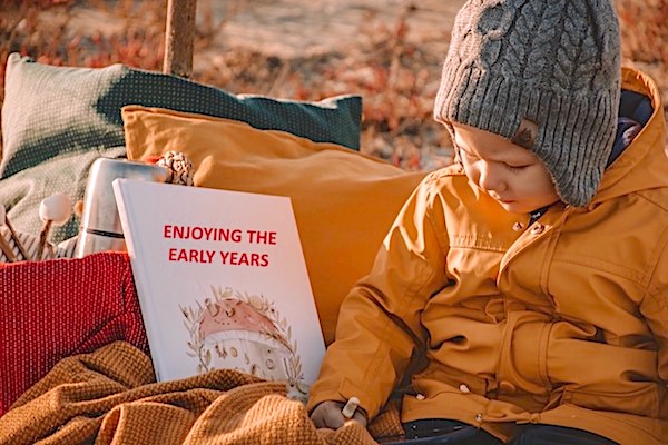 Young child wrapped up in warm coat and hat with a book nearby which says 'Enjoying the Early Years'