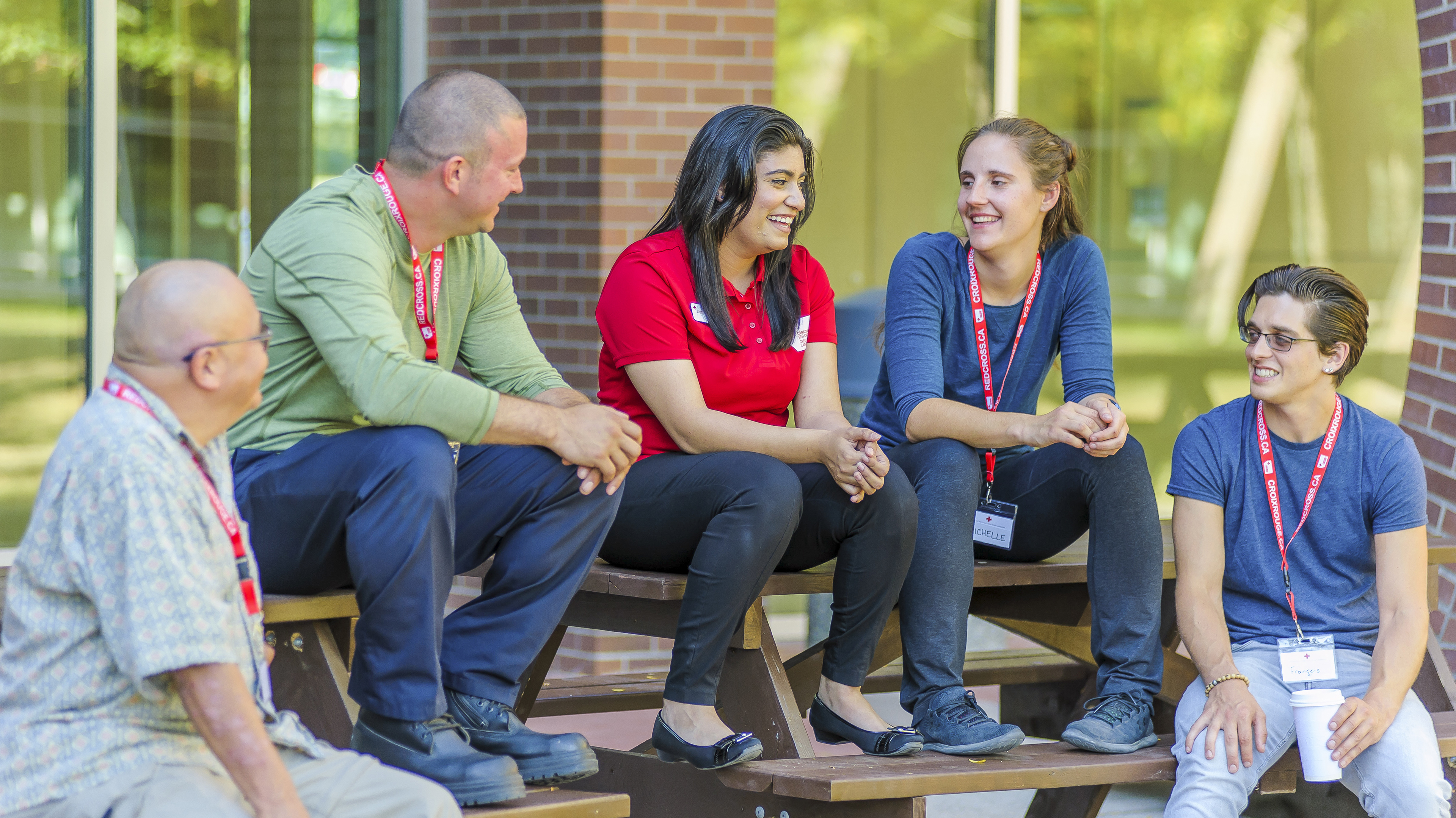 A group of people are gathered outside a building, chatting and enjoying each others company. Each person is wearing a Red Cross identification.