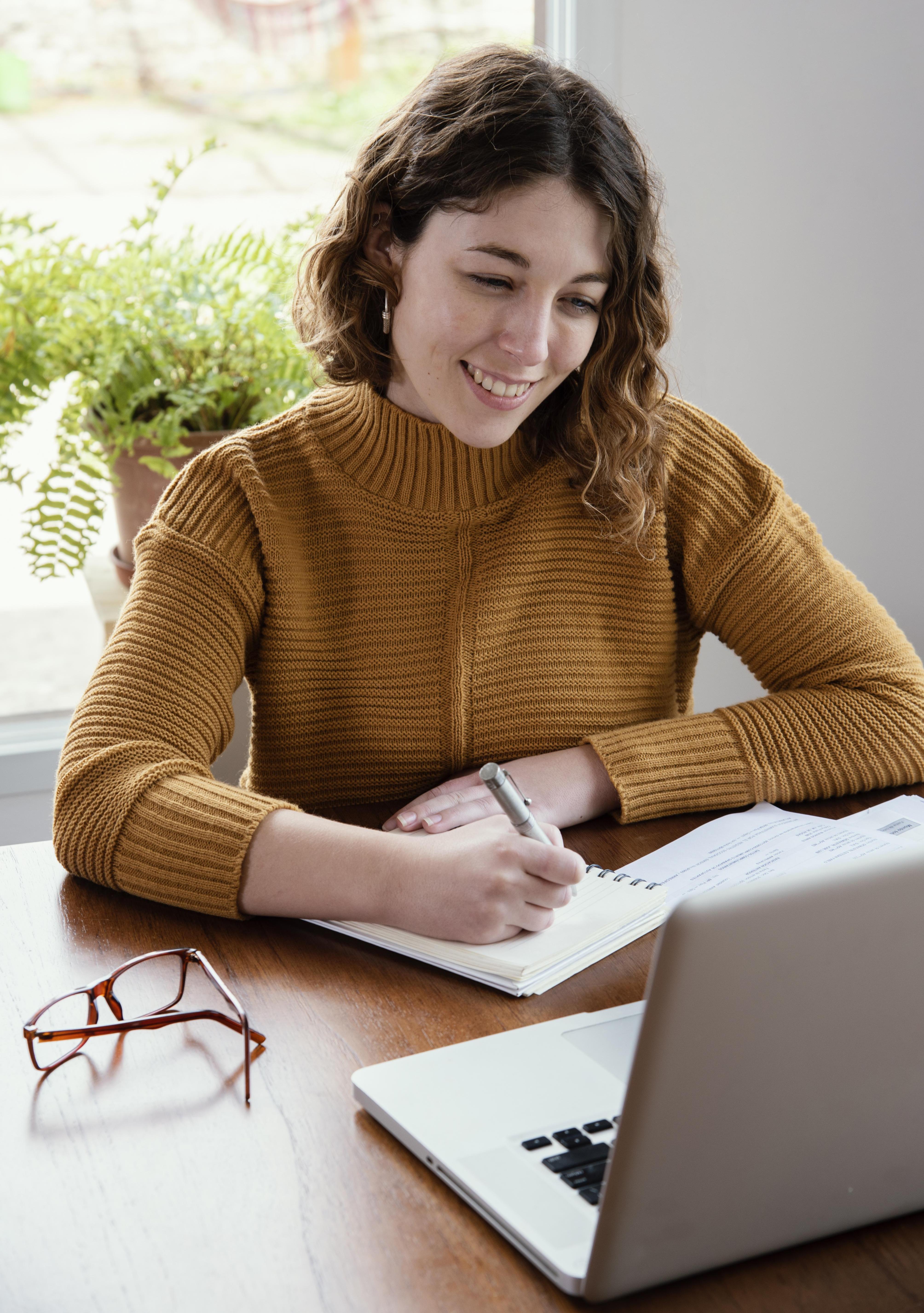 A woman at a desk taking an online course, she is smiling and making notes in a notebook 