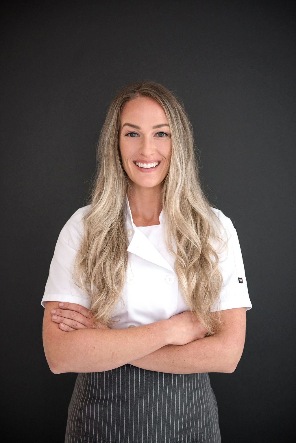 Chef Siiri Sampson stands in front of a black backdrop in her chefs coat and apron, arms folded and smiling.