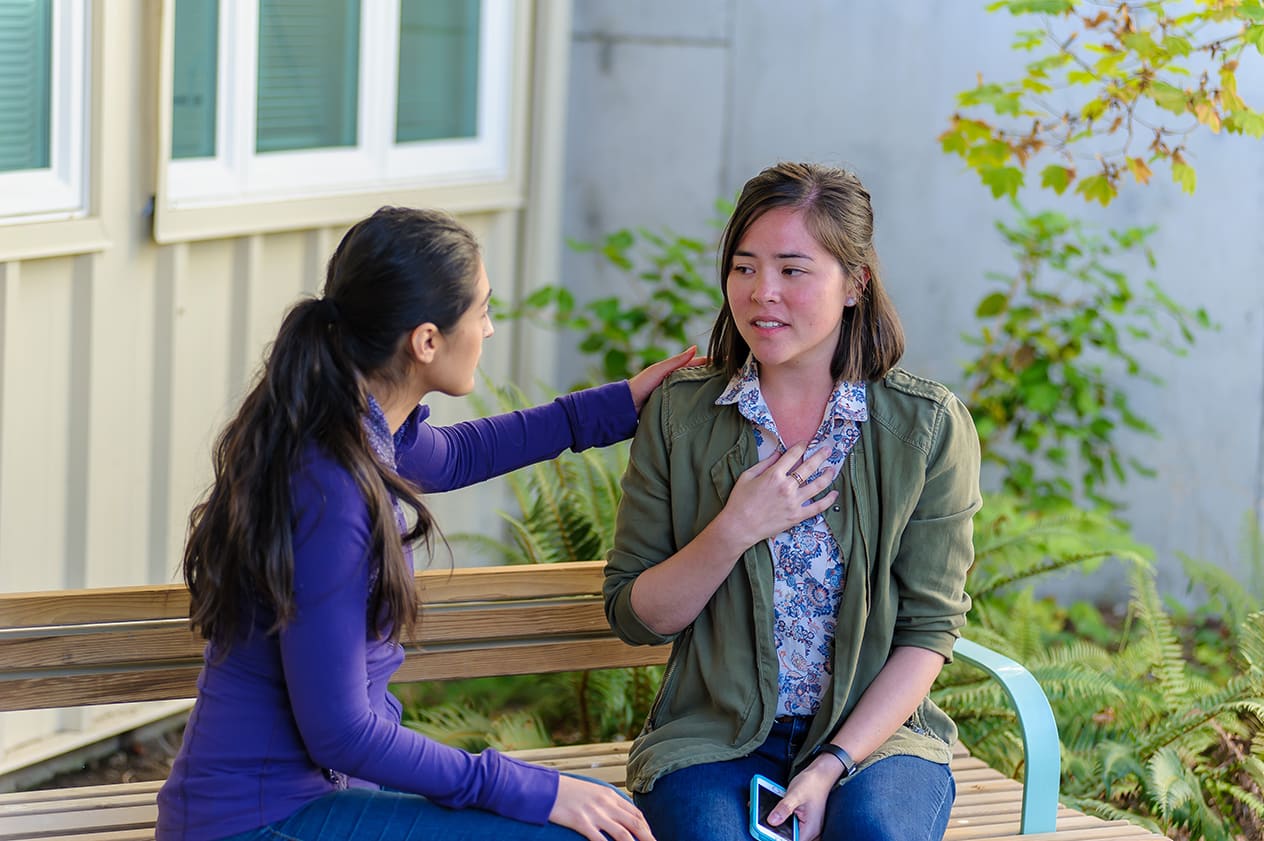 Une femme assise sur un banc tient sa poitrine et a de la difficulté à respirer alors qu’une autre femme s’approche d’elle.