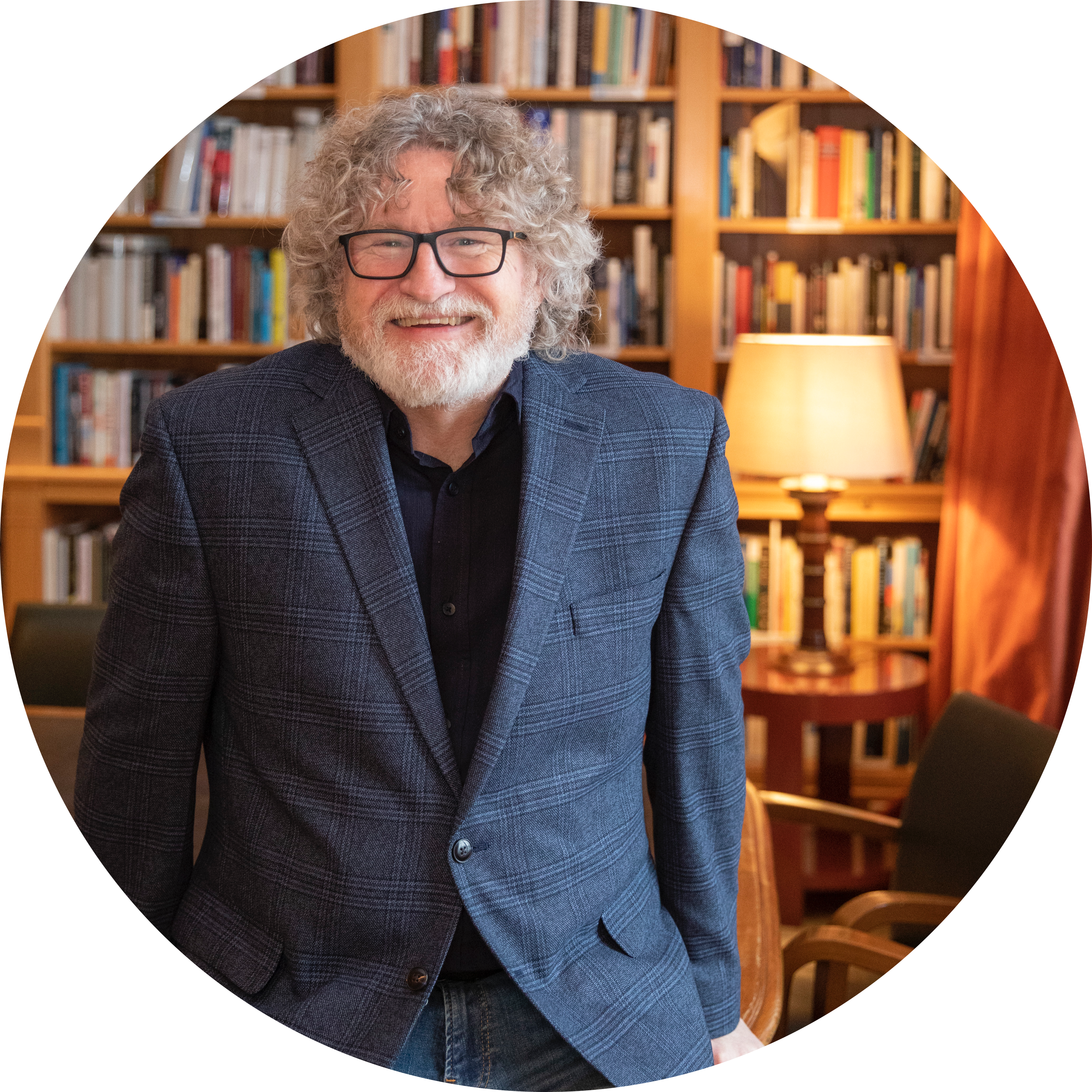 Smiling, author and teacher Brian Evenson wears a grey suit jacket, black shirt and black famed glasses standing in front of a bookshelf and lamp