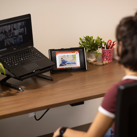 Une personne assise à un bureau suit la formation en ligne Secourisme en cas d’empoisonnement aux opioïdes à l’aide d’une tablette. Un ordinateur portable et des fournitures de bureau sont également visibles. 