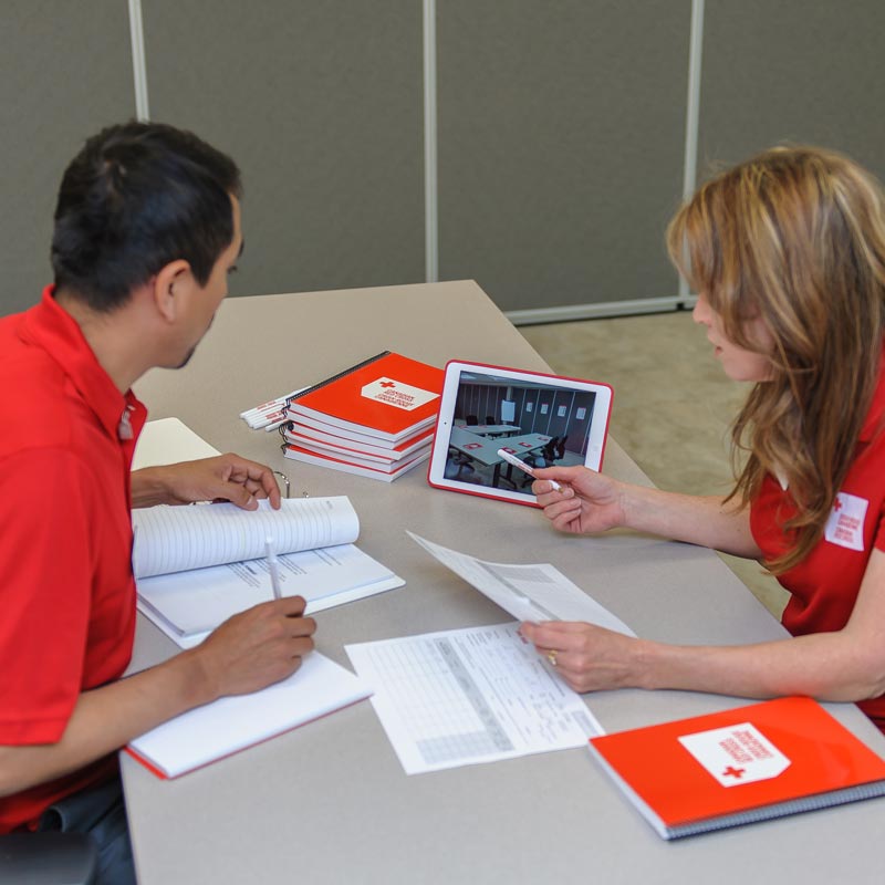 First Aid Instructor in a Red Cross shirt sitting at a table and looking at a tablet with a learner. 