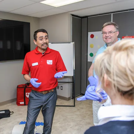 Person wearing a Red Cross Instructor shirt standing in a classroom speaking to two learners.