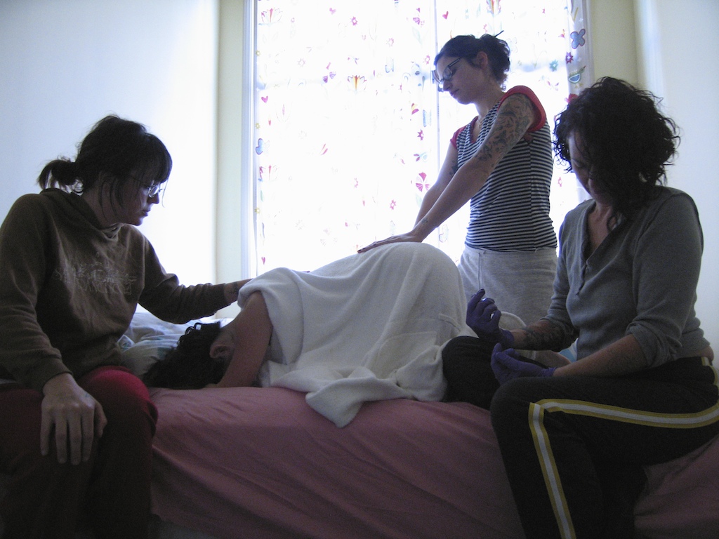 Two masc presenting people sit together shirtless, one holding their newborn baby skin to skin with a blanket over them, the other gazing lovingly at the baby over his partner&#39;s shoulder.