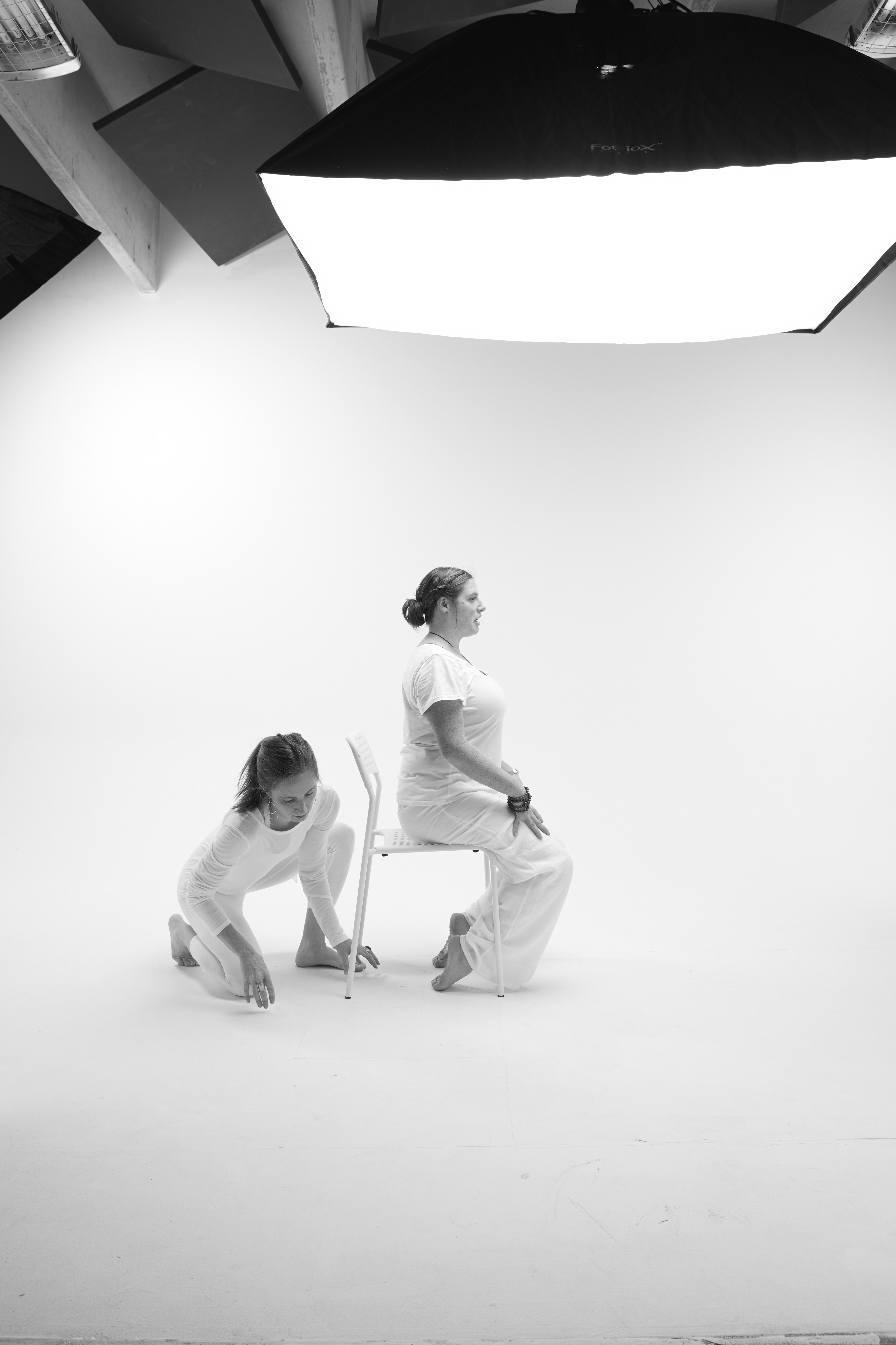 black and white image of yin yoga teacher Kaitlin Jackson demonstrating toe pose using a chair and Kari Kwinn preparing to support her