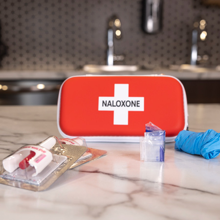 Une trousse de naloxone repose sur le comptoir d’une cuisine entourée de son contenu : deux doses de naloxone, une paire de gants sans latex et une barrière de protection respiratoire.