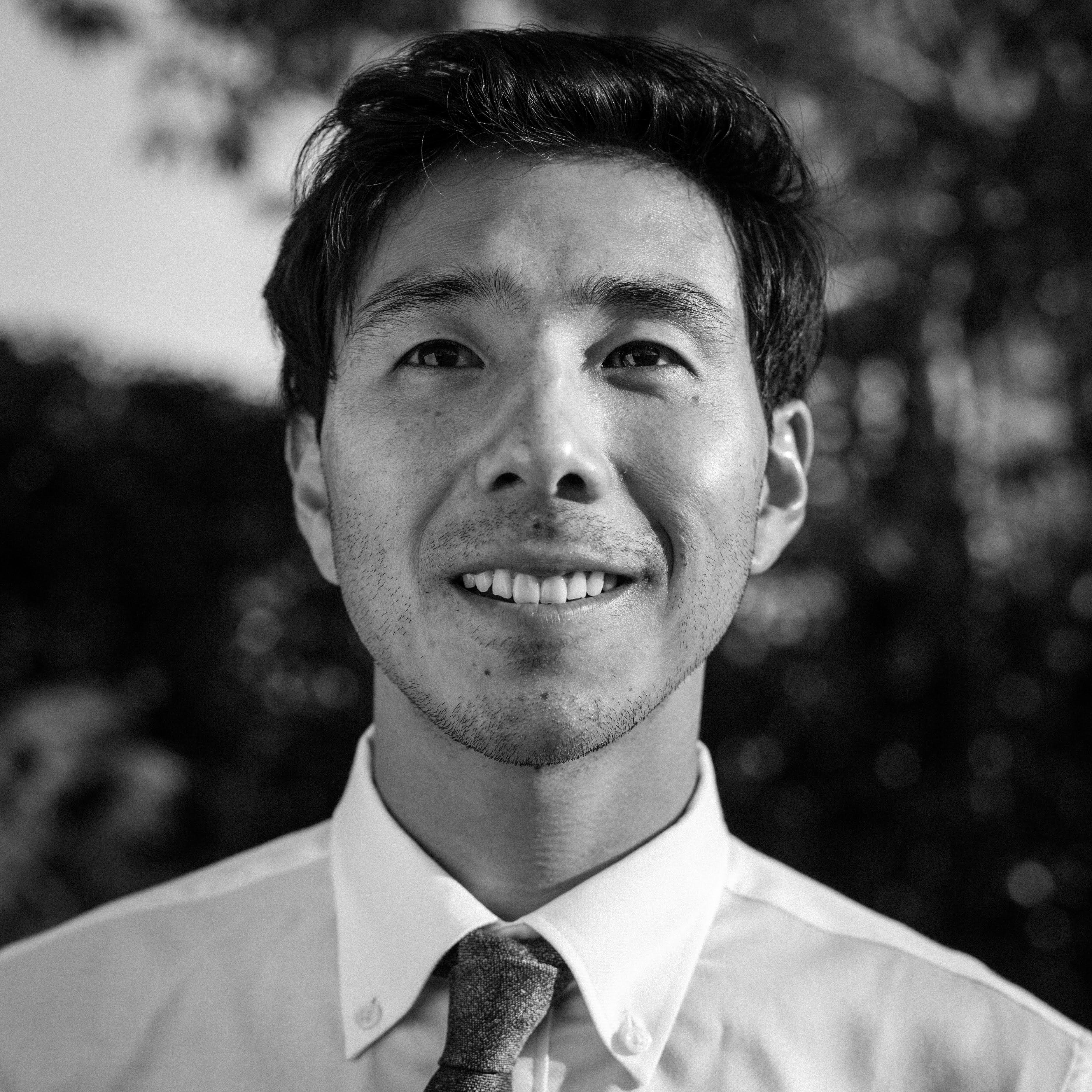Black and white headshot of Garrett Oyama, a Japanese-American man, smiling and looking up. He&#39;s wearing a white suit shirt and tie. There are trees in the background.