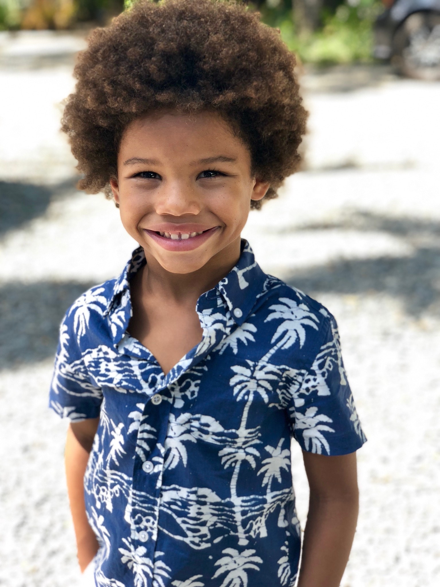 A 5 year old African American boy is standing in a blue and white Hawaiian shirt, smiling at the camera. 