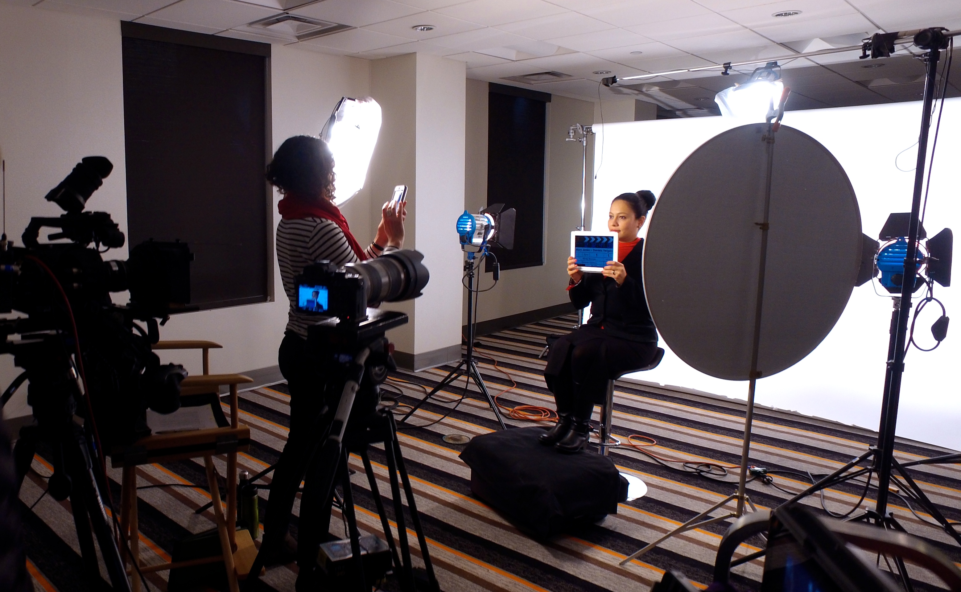 A female film director takes a photo of a dark-haired woman who is surrounded by cameras and lights on a film set