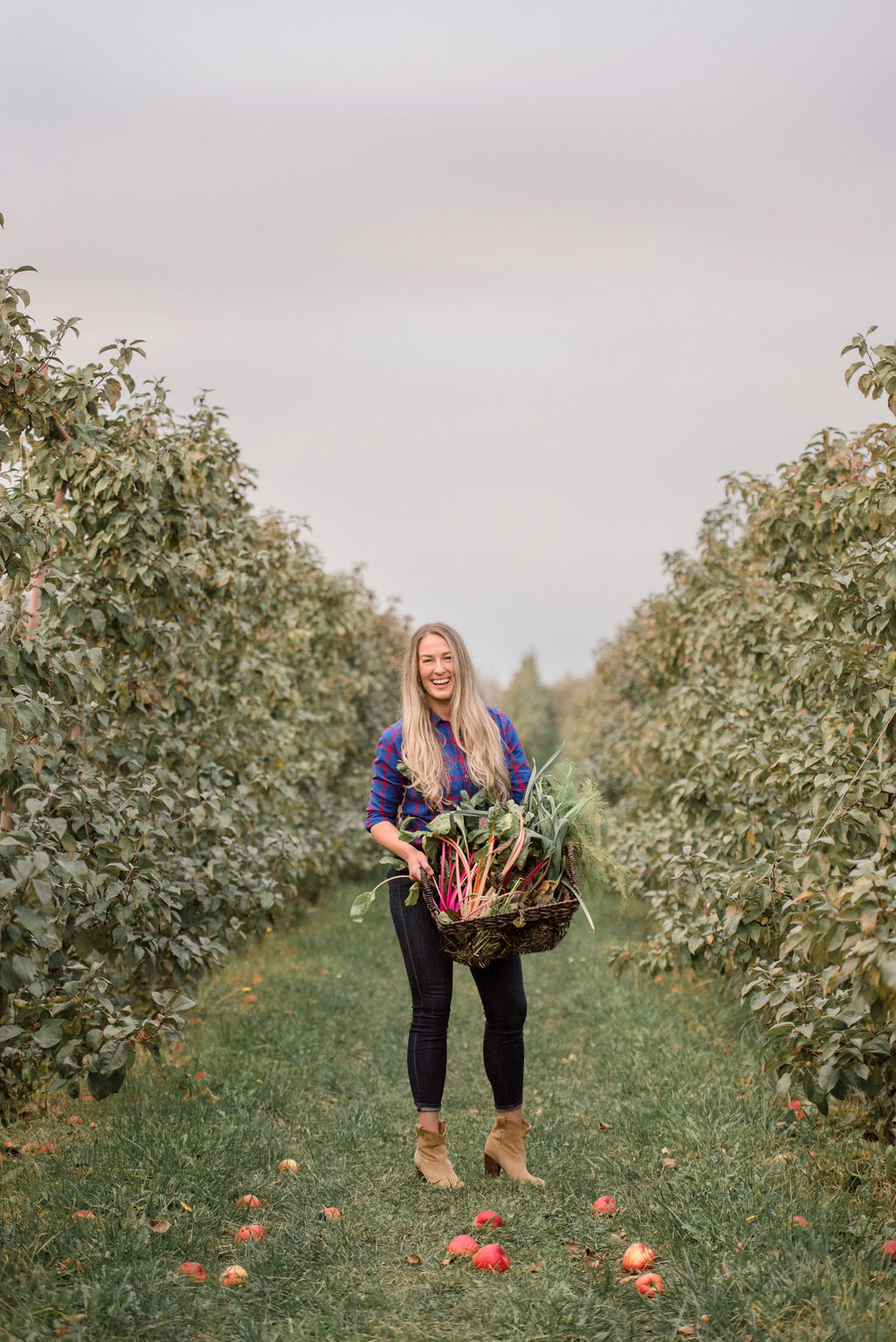 Chef Siiri Sampson stands in an apple orchard with a basket full of fresh produce grown from her garden.