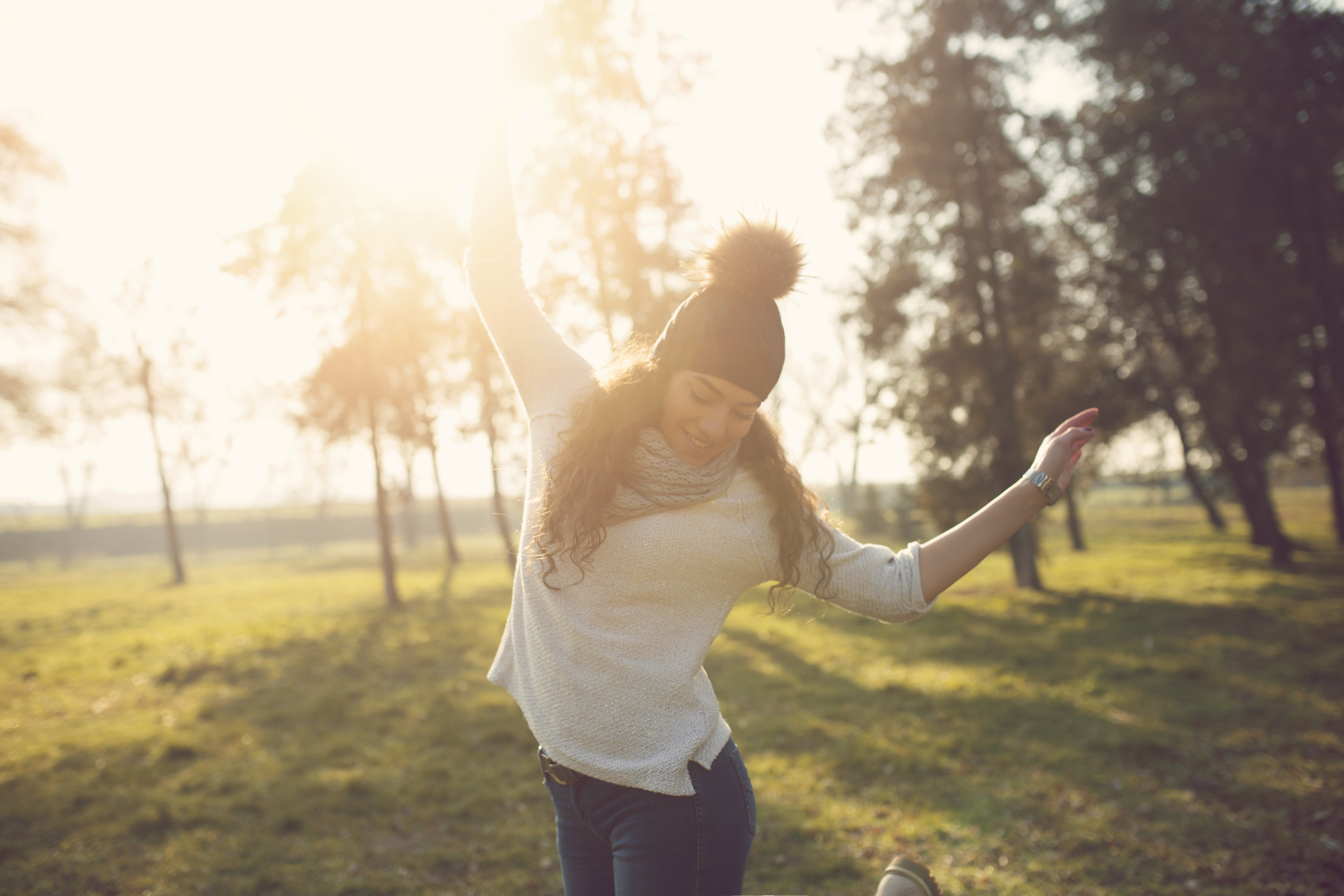 Long haired brunette woman wearing winter hat and dancing in a field with trees behind her