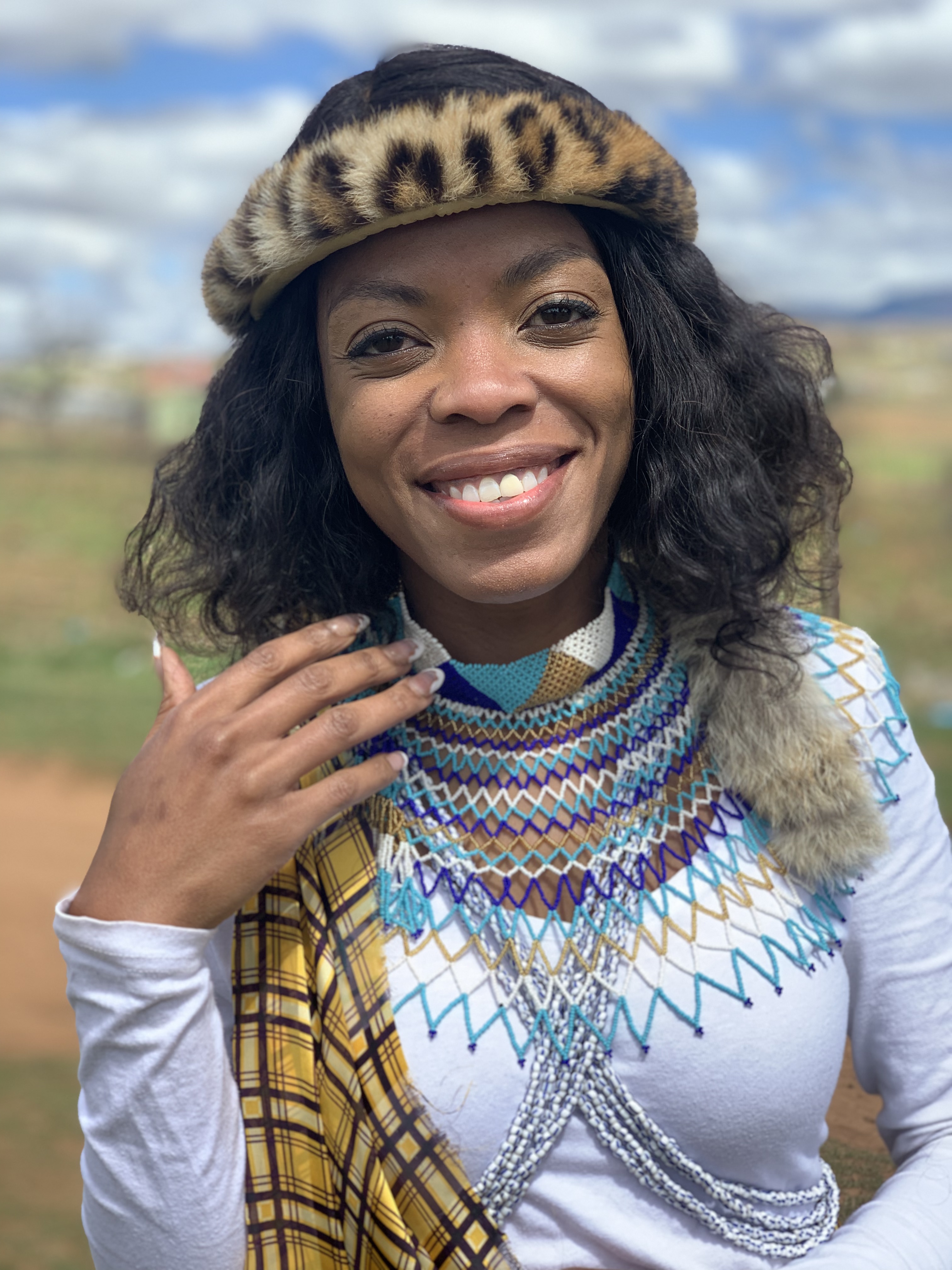 Makhosi Nomabutho looks into the camera. She wears a leopard headpiece, blue, gold and white beaded neckpiece as well as blue and white bead across her body over a white shirt, draped with a gold and purple ceremonial cloth. 