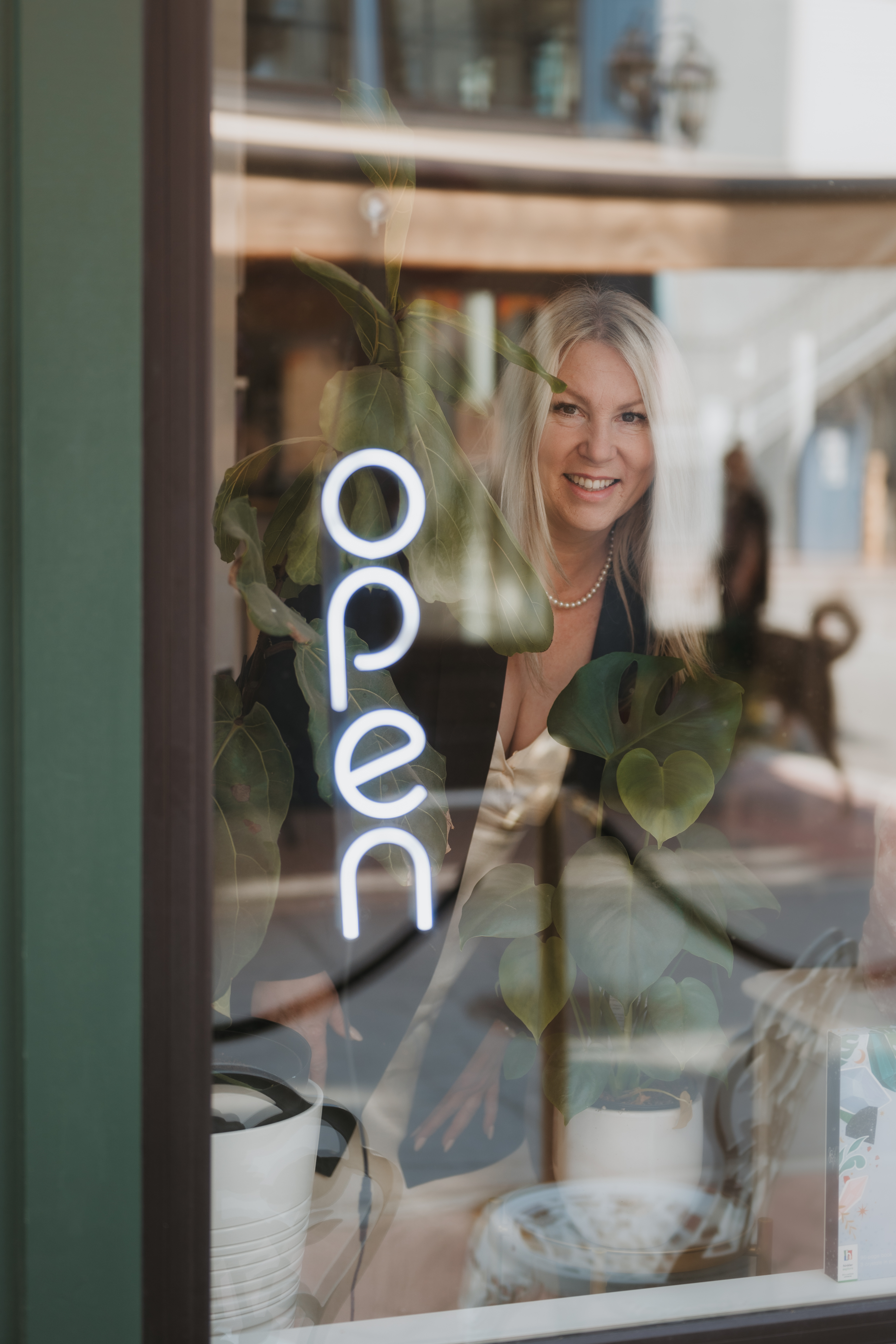 a smiling blond woman peering out a window between plants leaves and beside a white open sign