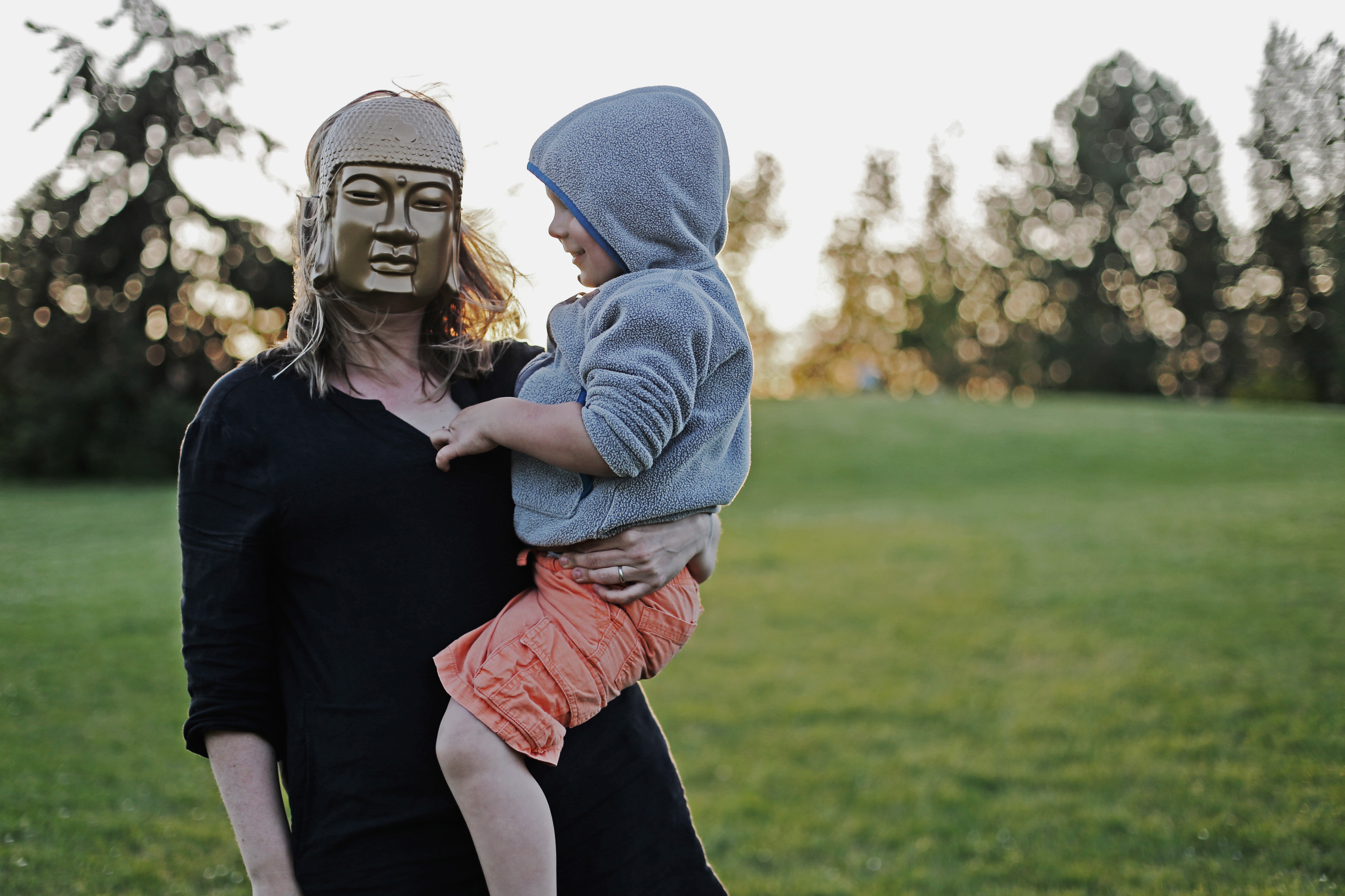 A woman wearing a Buddha mask holds a child on her hip.