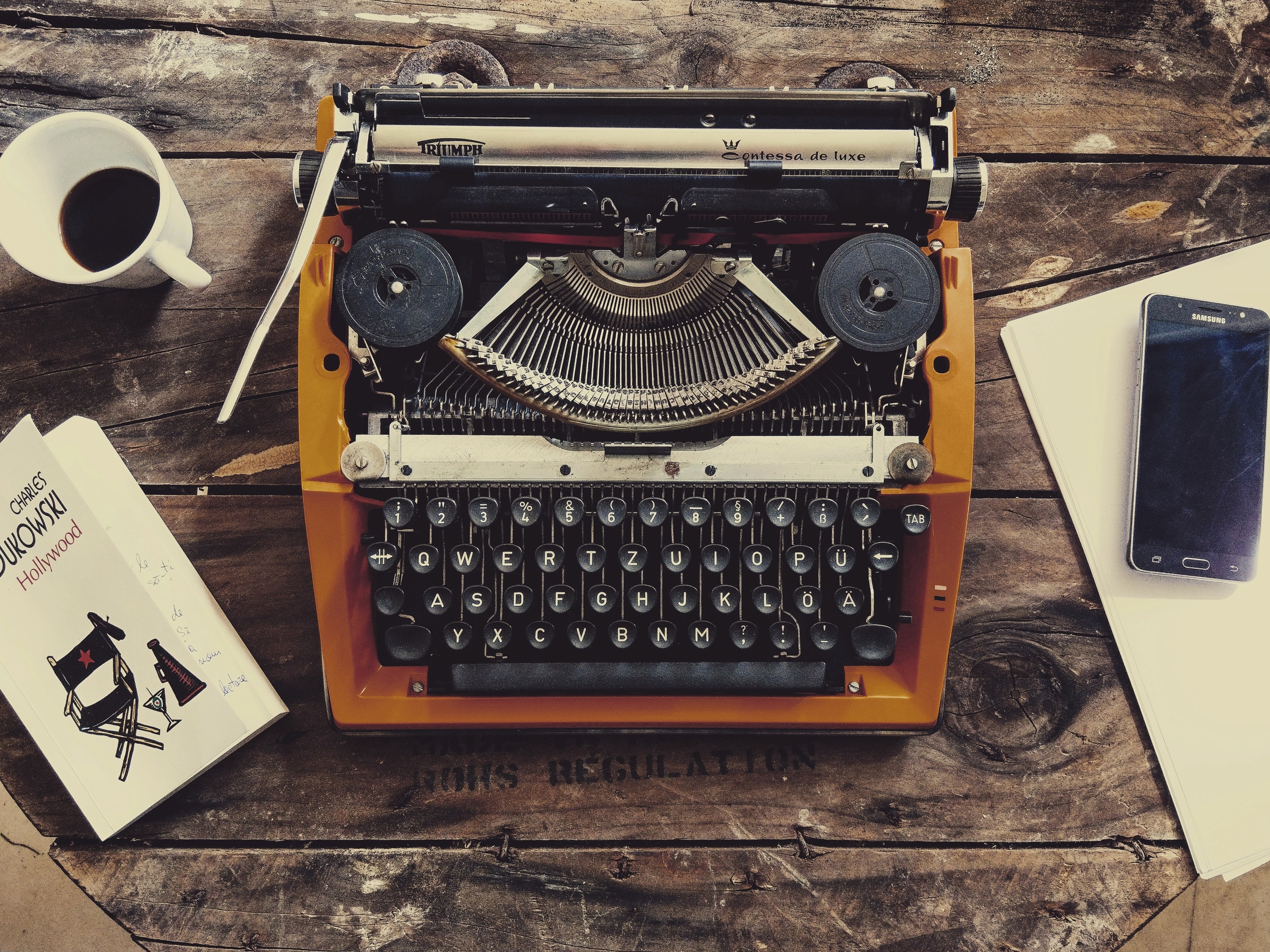 A classic typewriter rests on a wooden table, surrounded by coffee, papers, and a book.