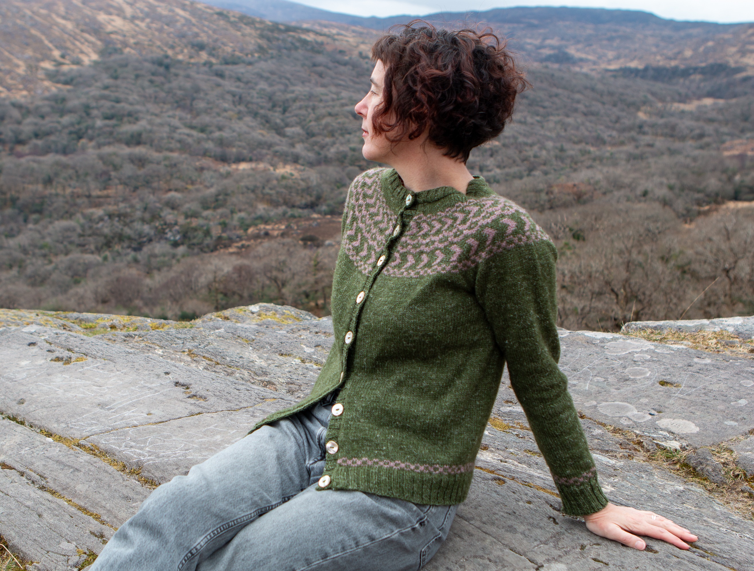 Carol Feller wearing the Daingean Cardigan sitting on a rock, with the view of a mountain range behind her. 