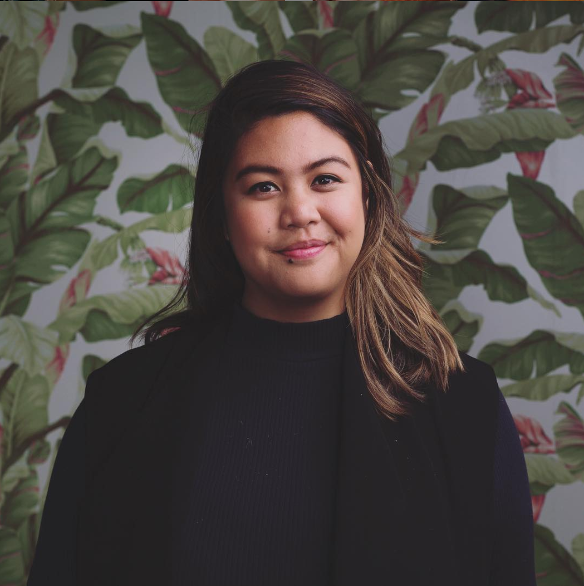 Smiling, young, Asian-American woman with medium-length brown hair with blonde highlights in a black turtleneck sweater. Background is white wallpaper patterned with pink flowers and green leaves.