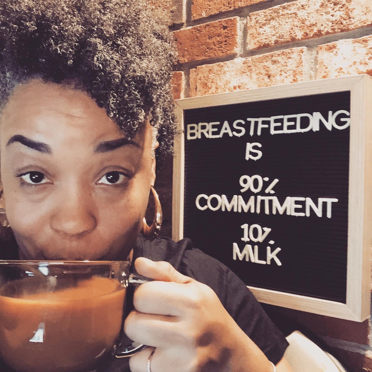 Light skinned Black woman holding a cup of coffee looking into the camera with a letterboard behind her that says breastfeedin 90 percent committment and 10 pecent milk.
